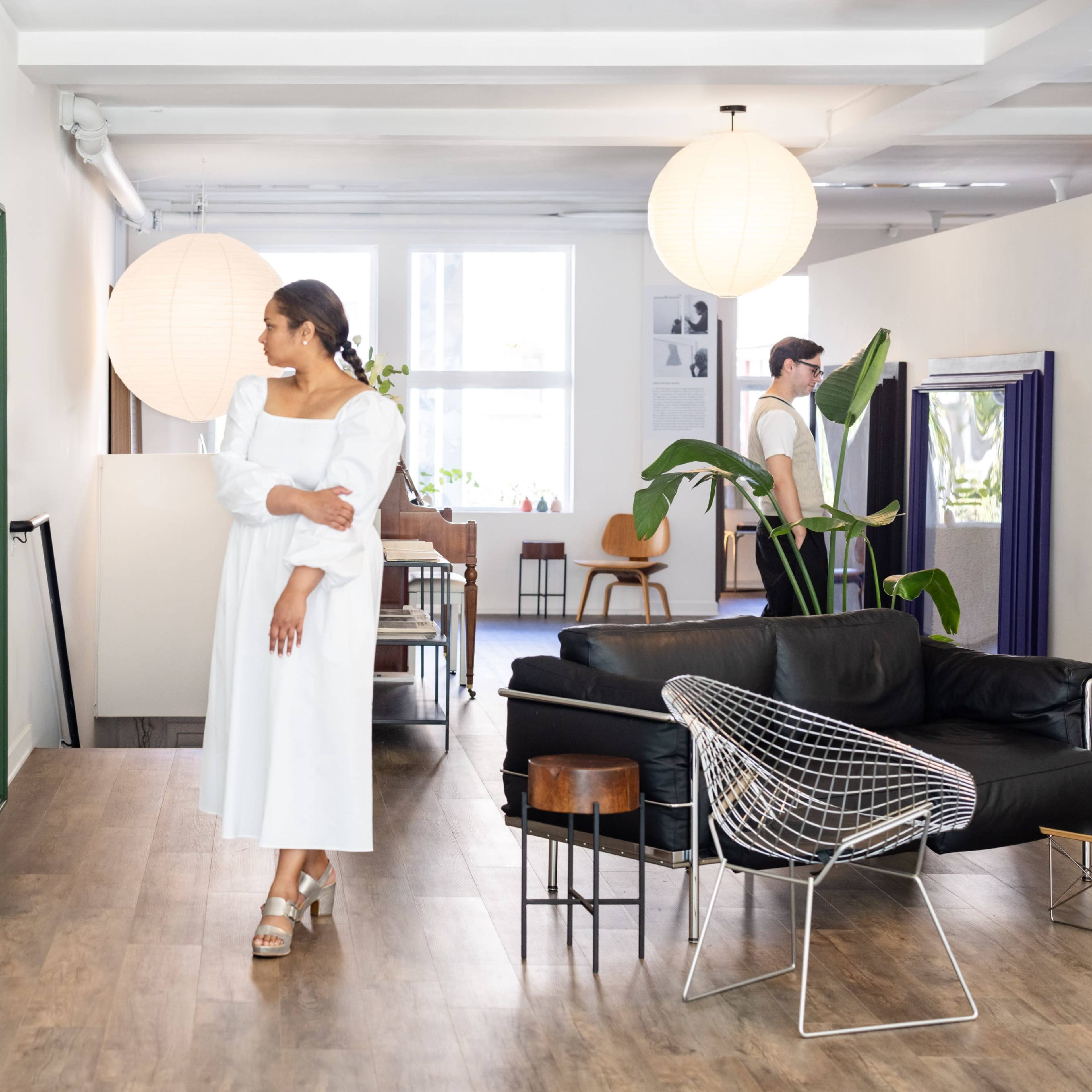 A woman in a white dress stands in a bright, modern interior featuring a mix of furniture and decorative plants.