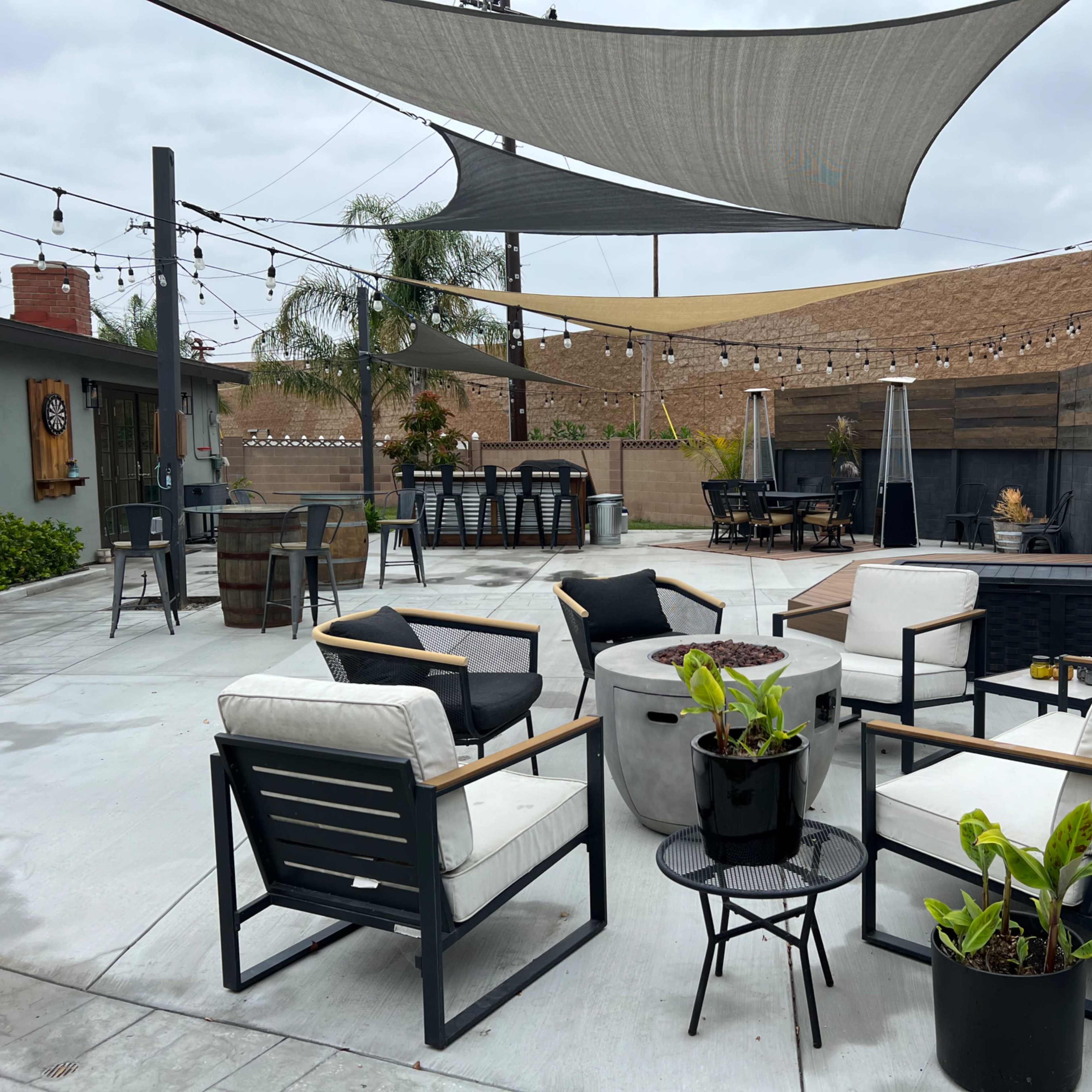 An outdoor patio area with modern seating, a fire pit, and shaded sections, surrounded by greenery and wooden fencing.