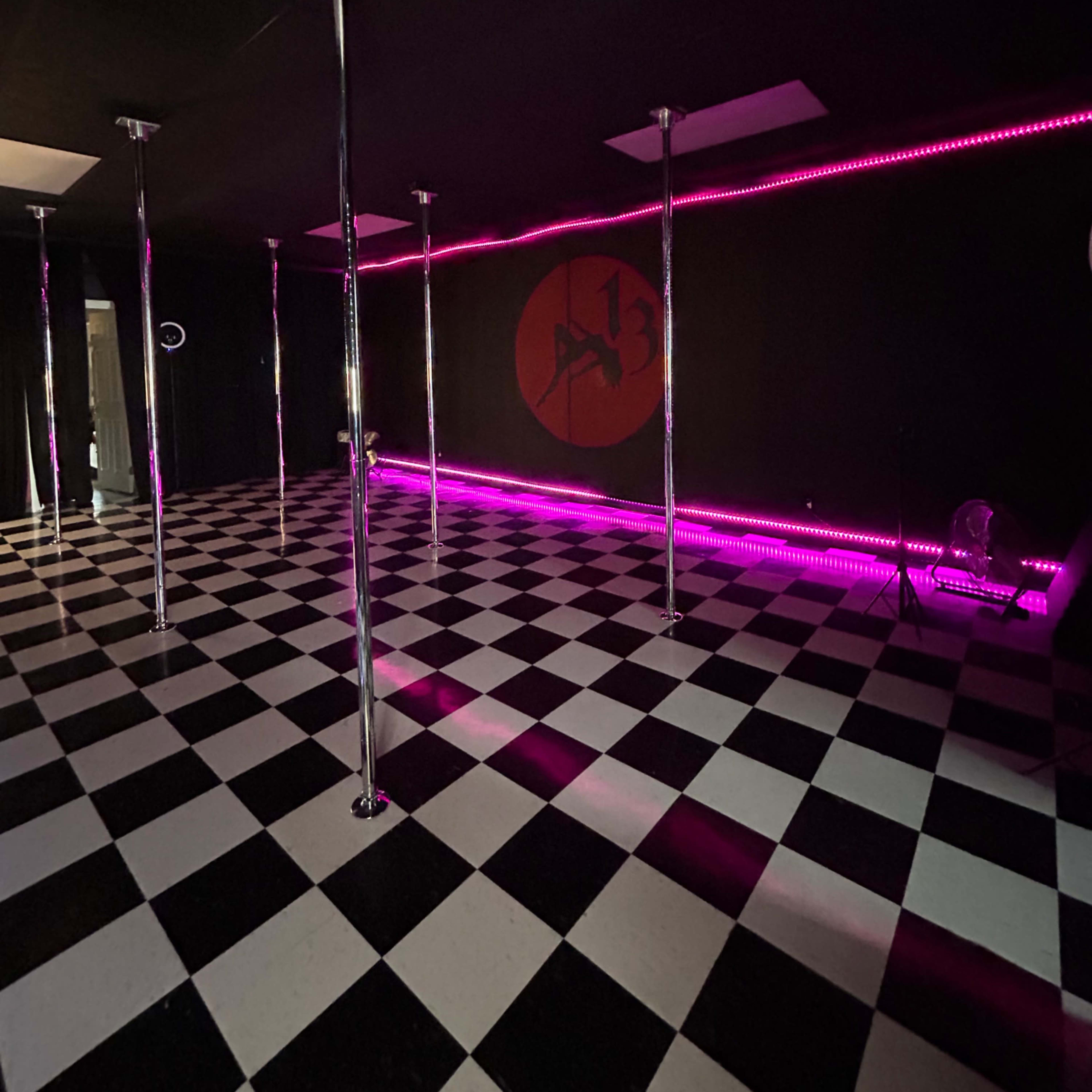 A room with black and white checkered flooring, several shiny poles, and pink LED lighting along the walls.