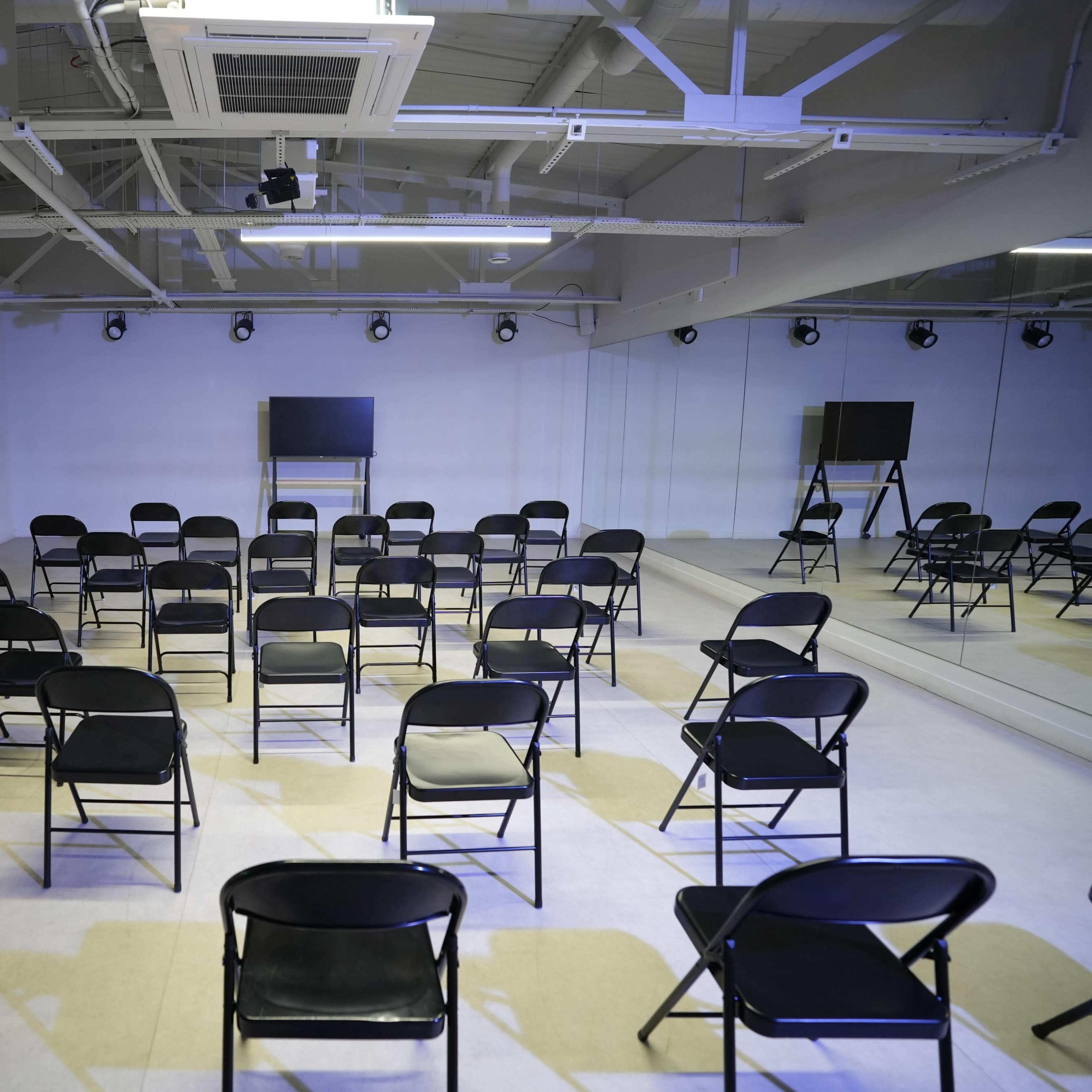 A room with a circular arrangement of black folding chairs facing a television screen, surrounded by mirrors.