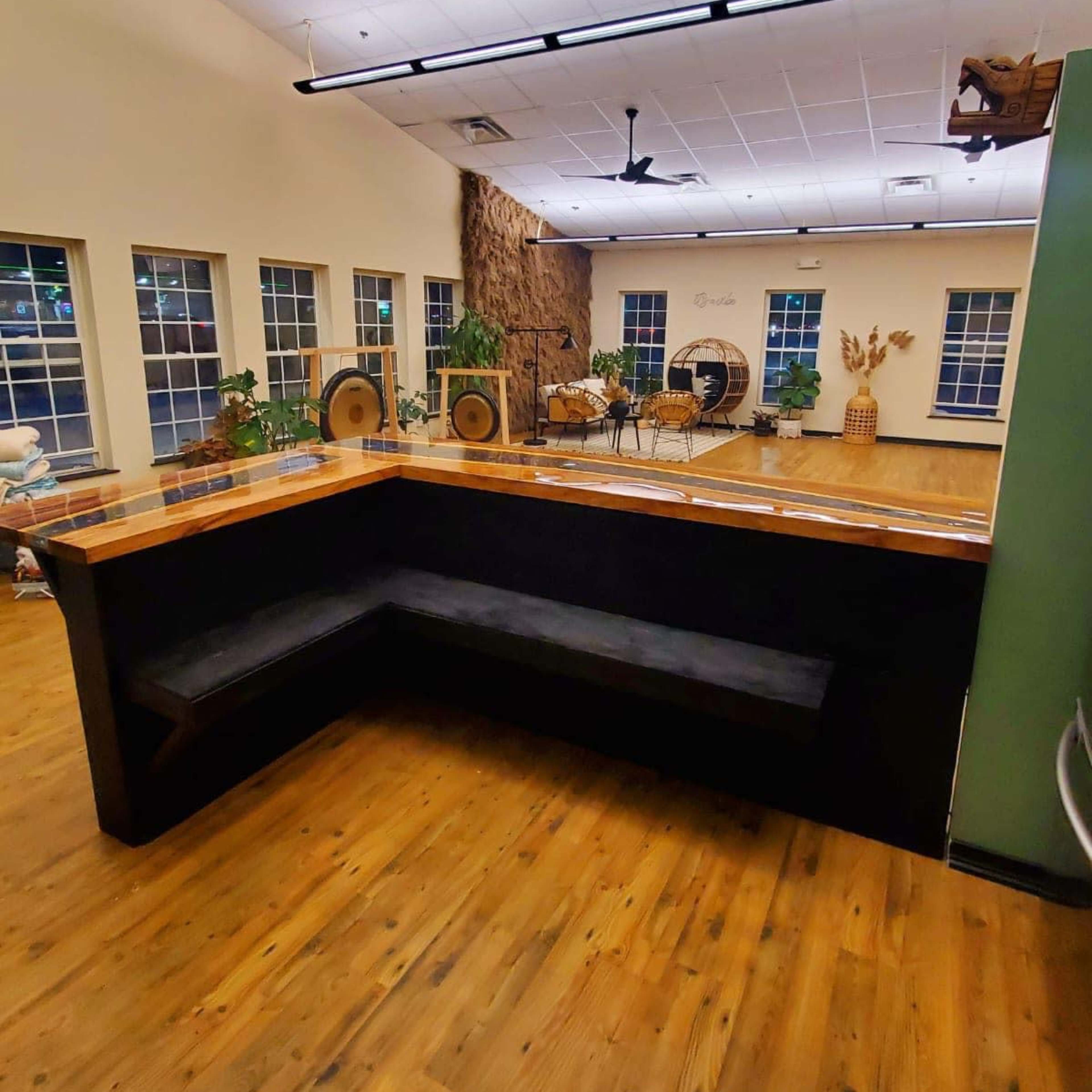 A spacious room with wooden flooring, featuring a black seating area against the wall and large windows allowing natural light.