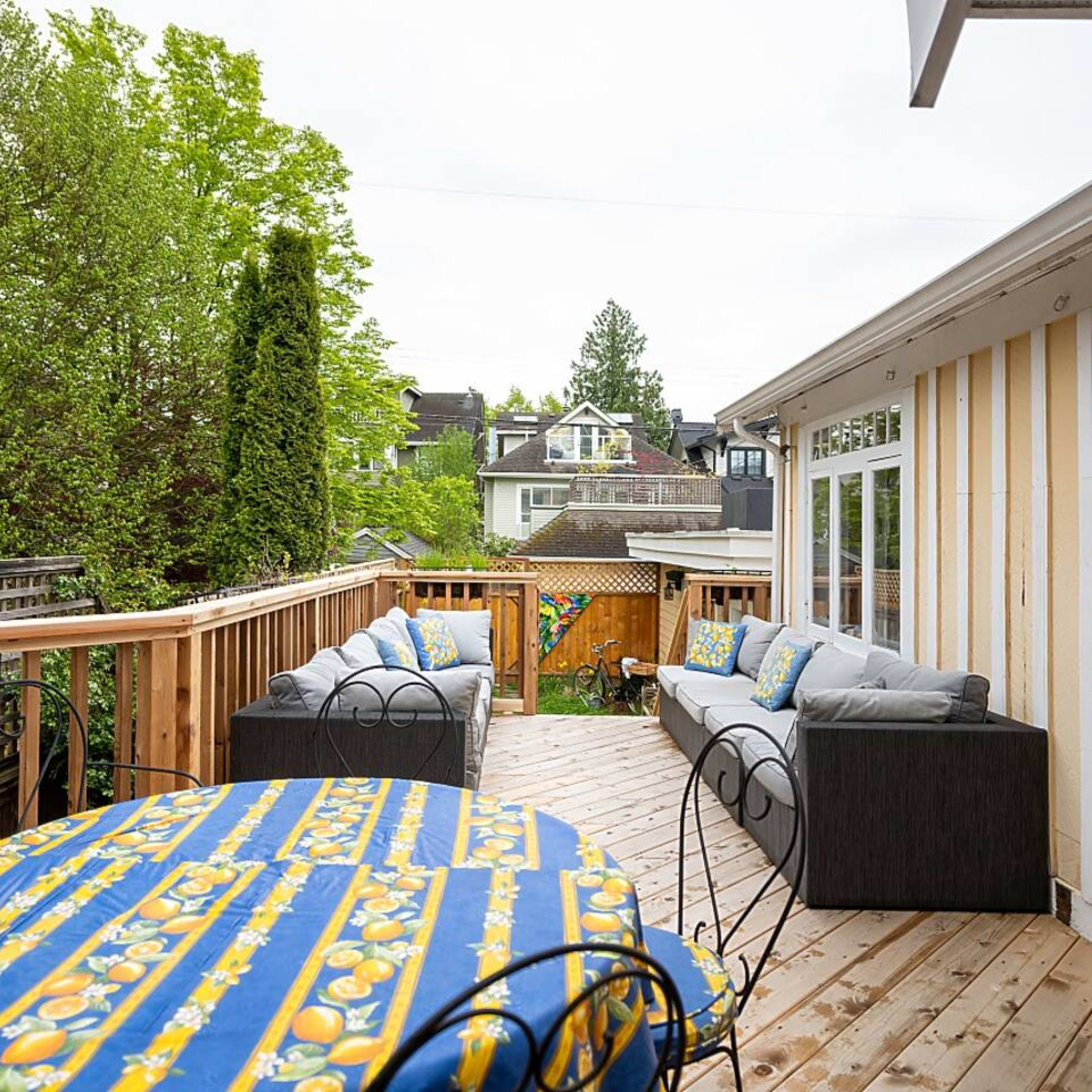 A wooden deck with a patterned tablecloth-covered table and a seating area furnished with gray couches, surrounded by greenery.