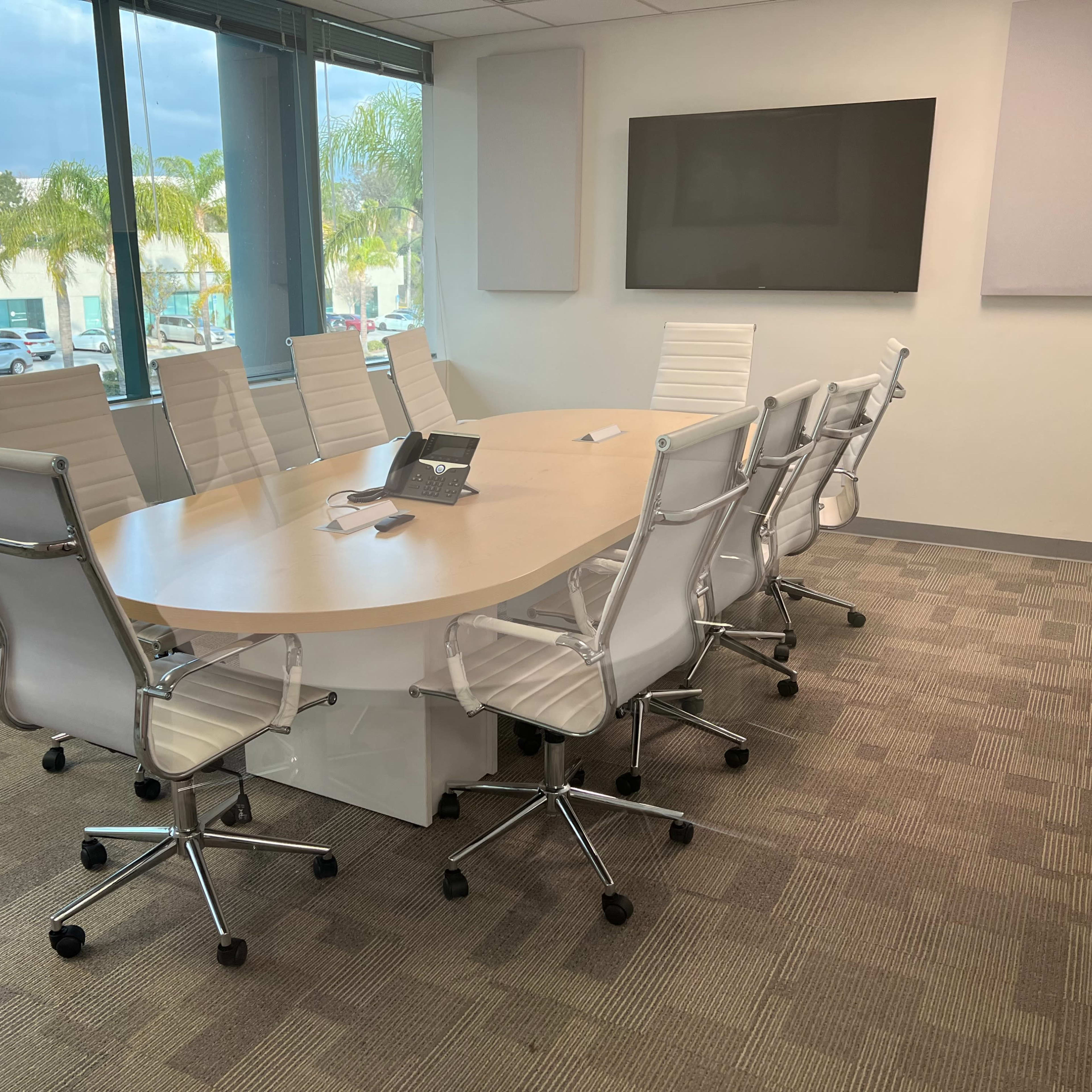 A modern conference room features a large oval table surrounded by white ergonomic chairs and a wall-mounted screen.