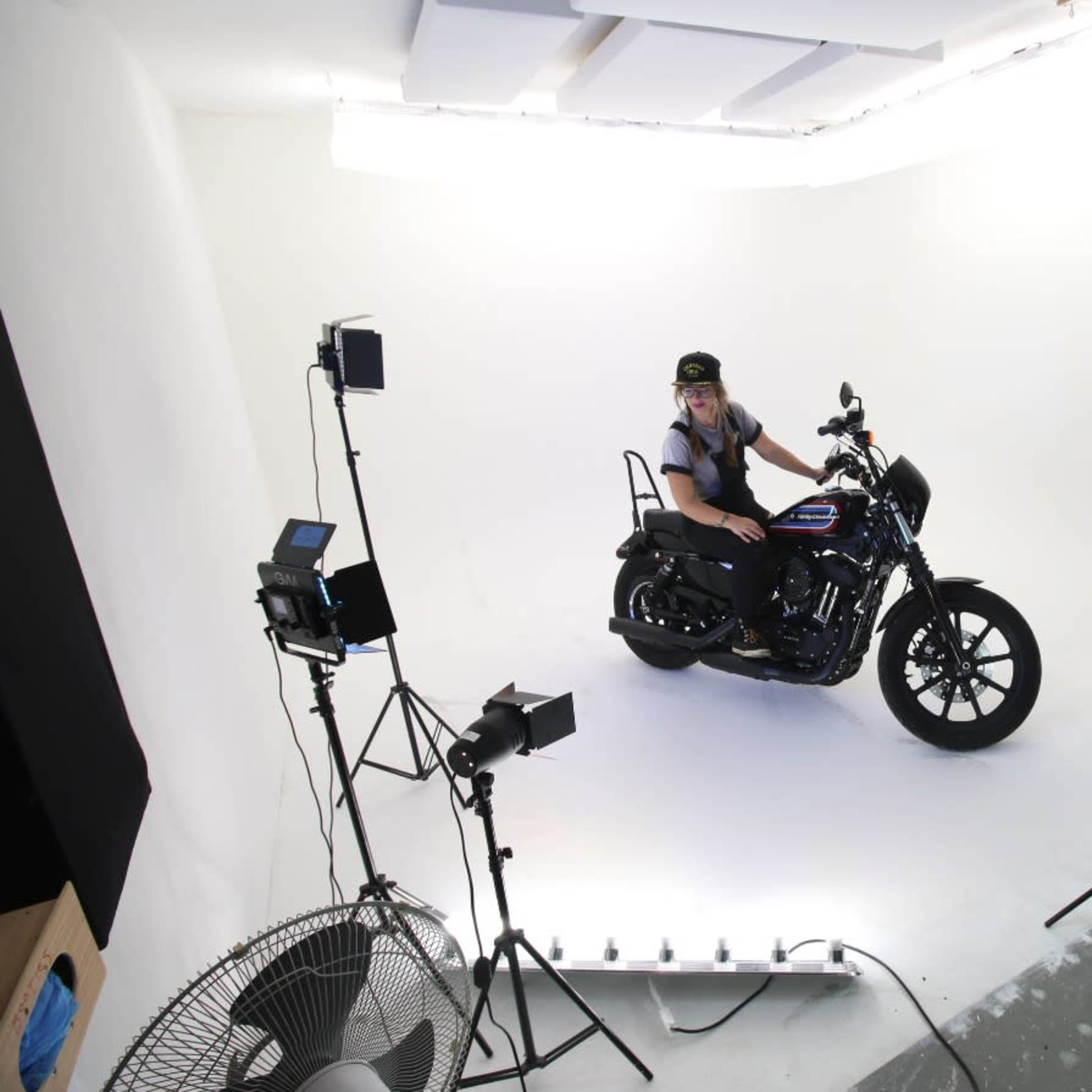 A person sits on a motorcycle in a brightly lit photo studio surrounded by lighting equipment.