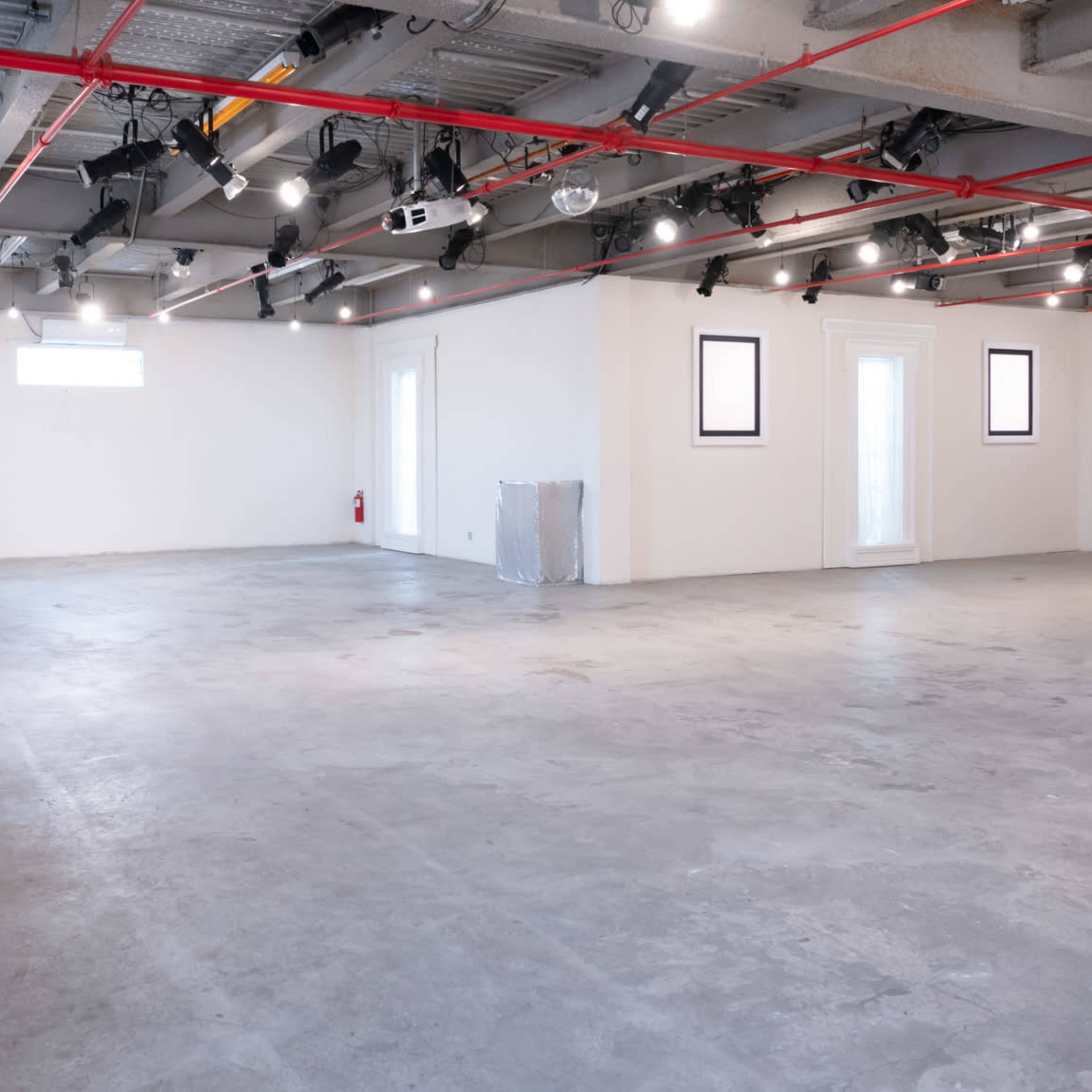 A spacious, empty room with concrete flooring, overhead lights, a ladder, and a few artworks on the walls.