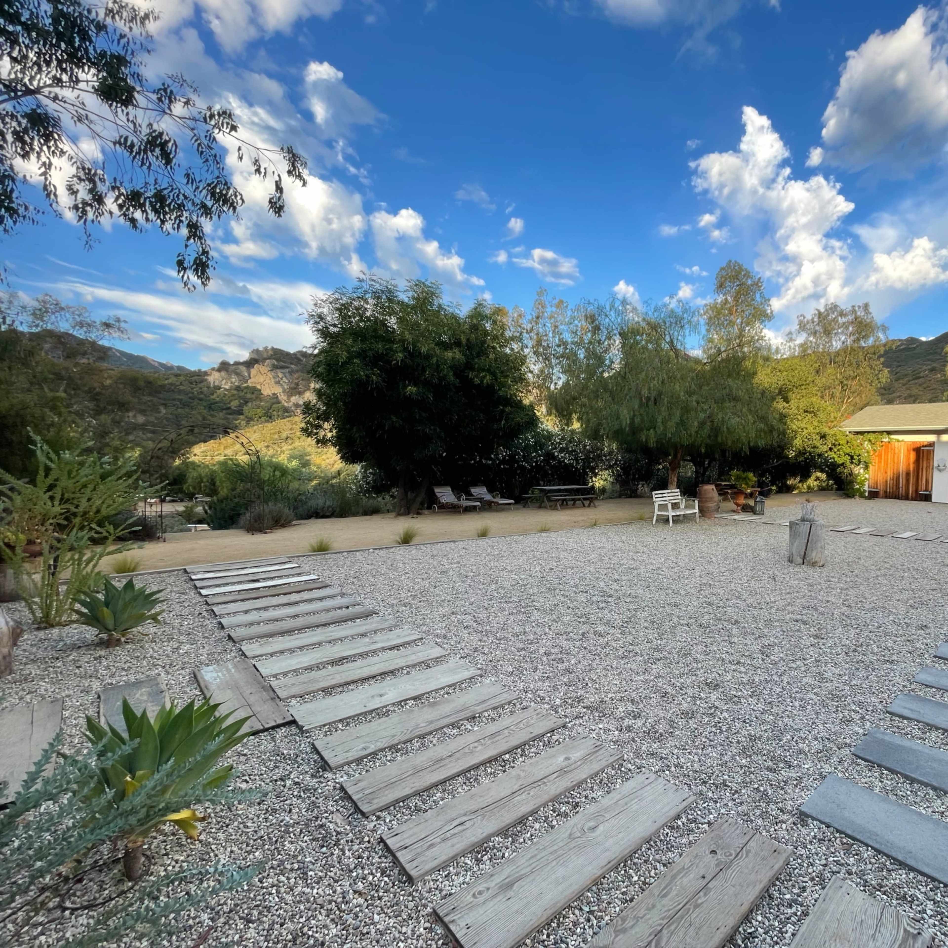 A gravel courtyard features wooden pathways leading to a white building, surrounded by greenery and mountains under a blue sky with scattered clouds.