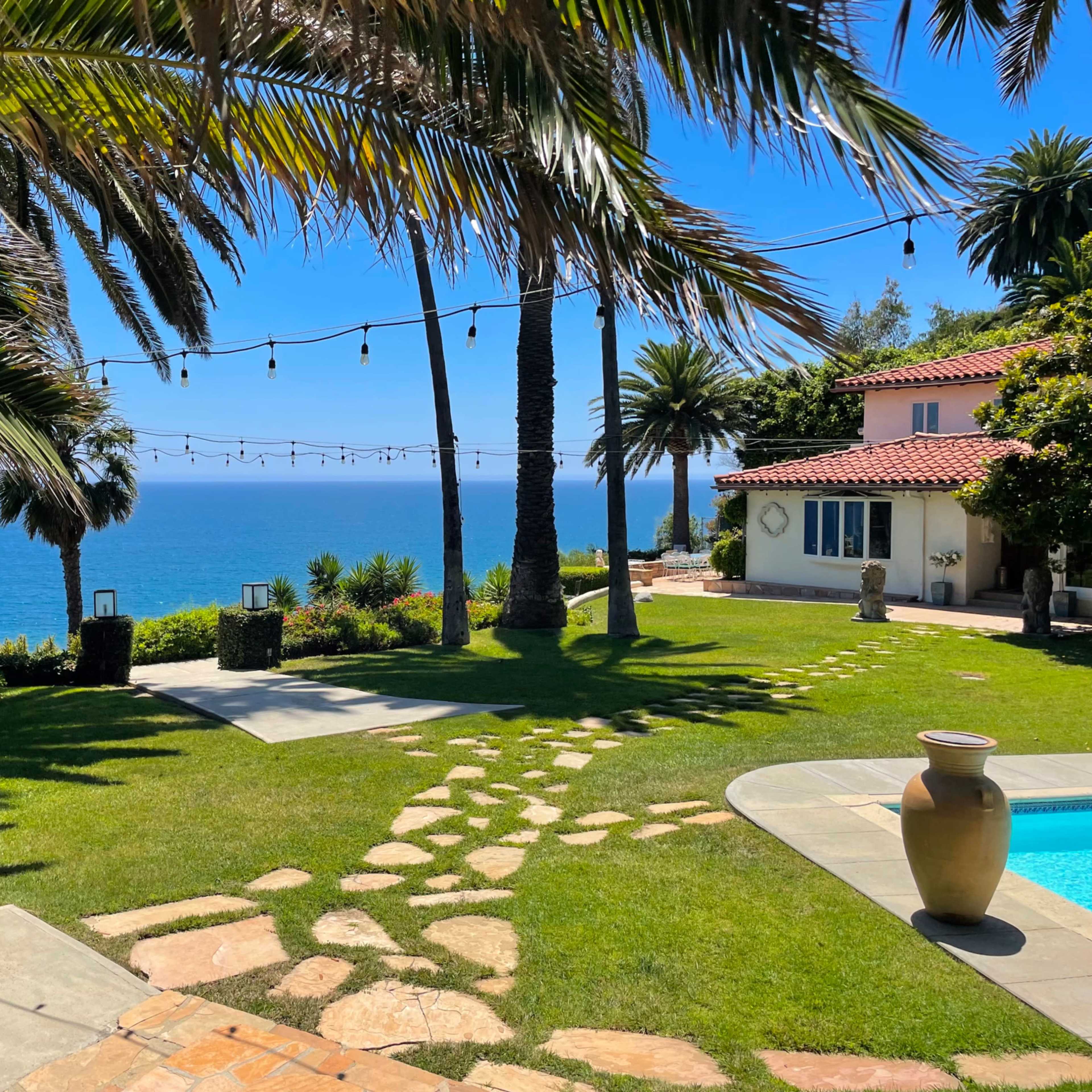 A landscaped yard features a stone pathway leading to a pool, with palm trees and ocean views in the background.