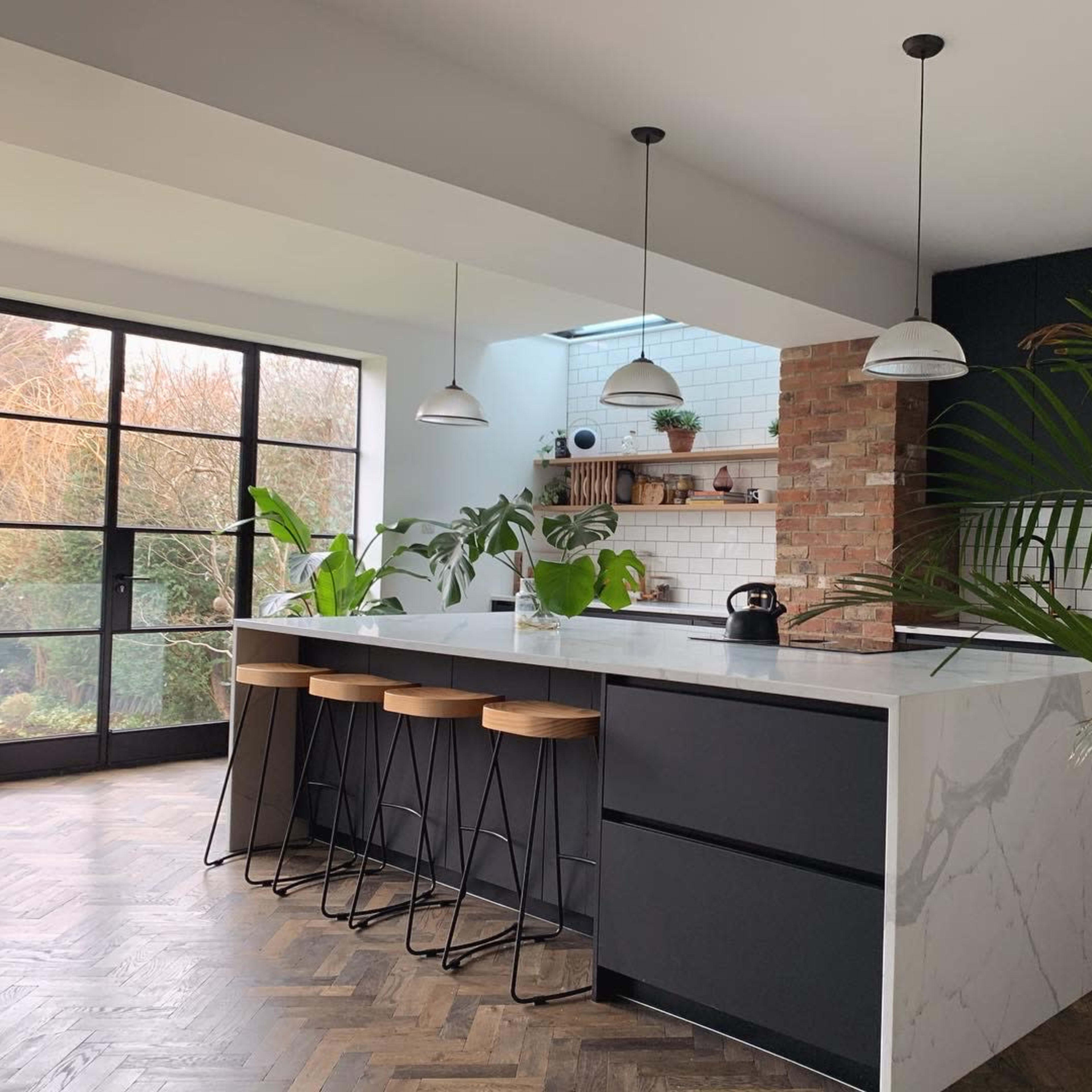 A modern kitchen with a large island, bar stools, and plants, featuring a mix of dark cabinetry and a light marble countertop.