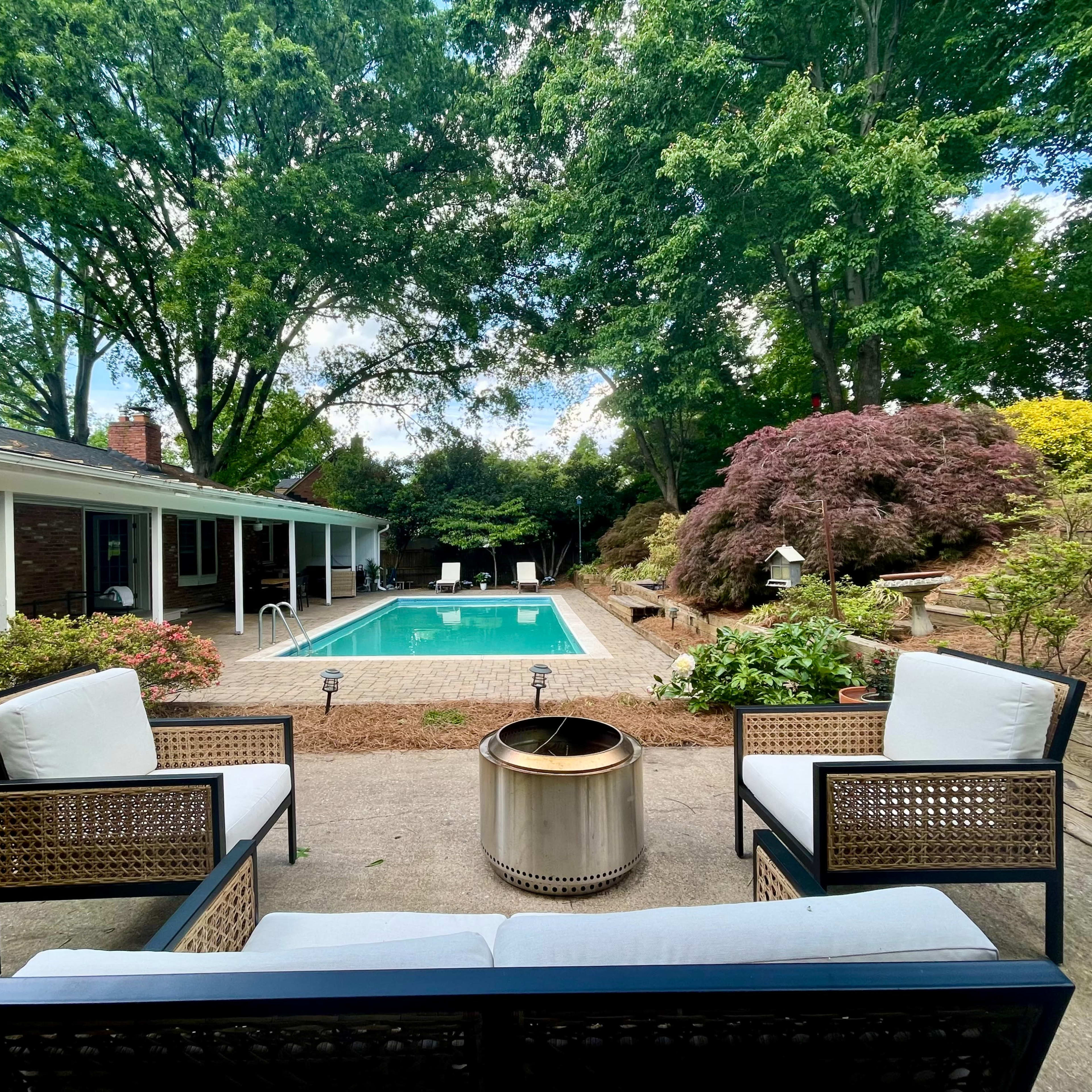 A patio featuring two chairs and a fire pit overlooks a swimming pool surrounded by landscaped greenery.