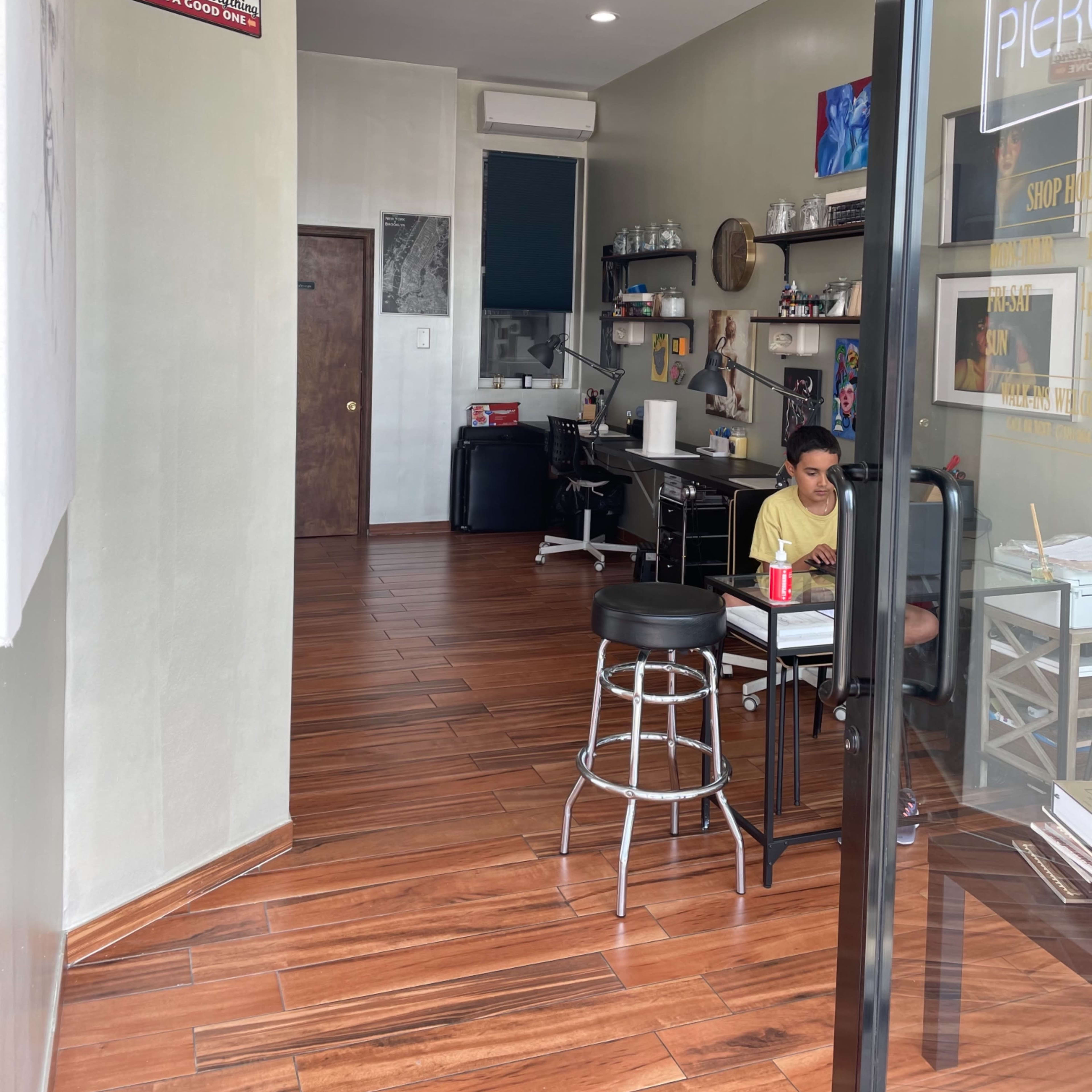The interior of a tattoo studio, featuring a workspace with a stool, desk, and various art supplies against a light-colored wall and wooden flooring.