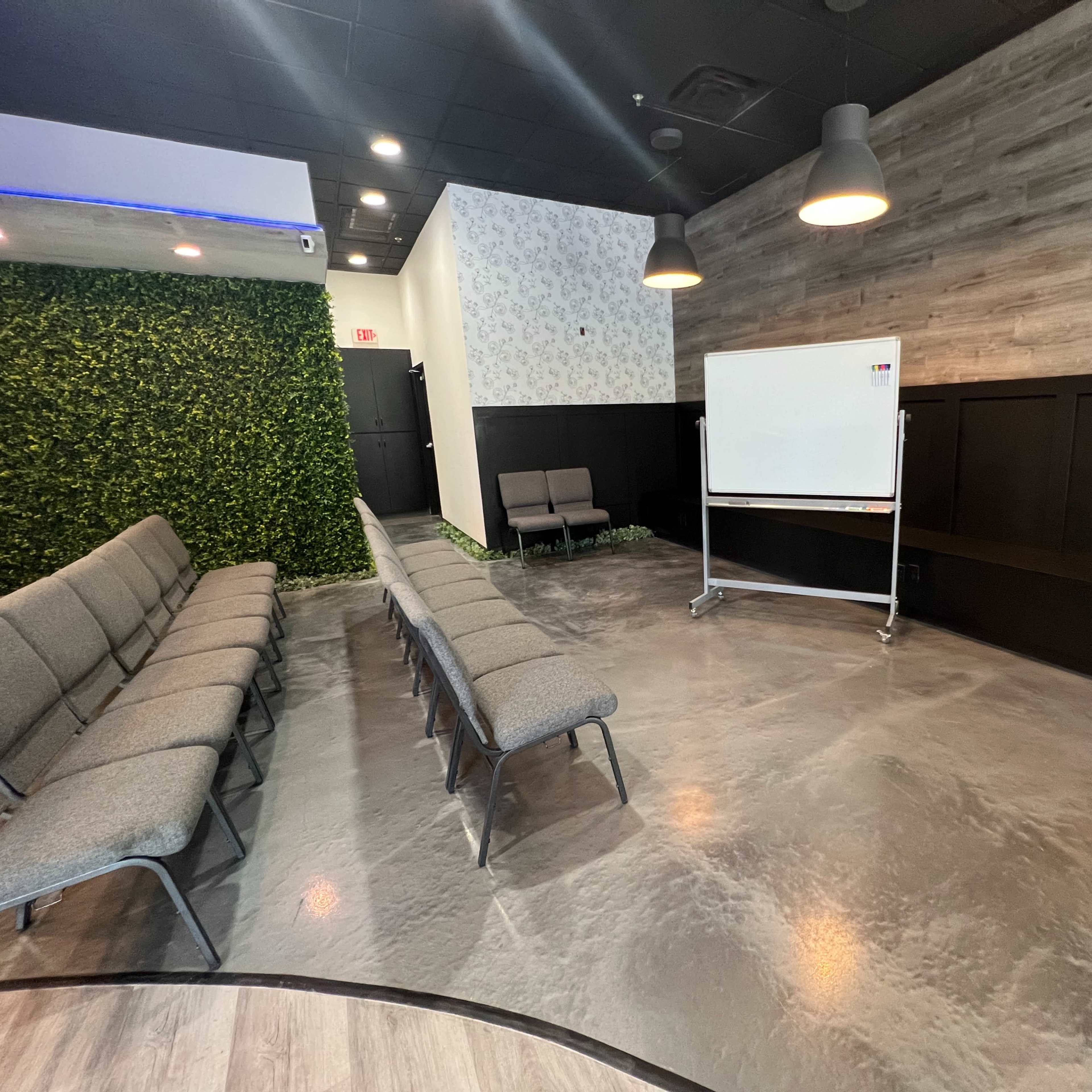 A modern meeting room with gray seating arranged in rows, a whiteboard on a mobile stand, and greenery along one wall.
