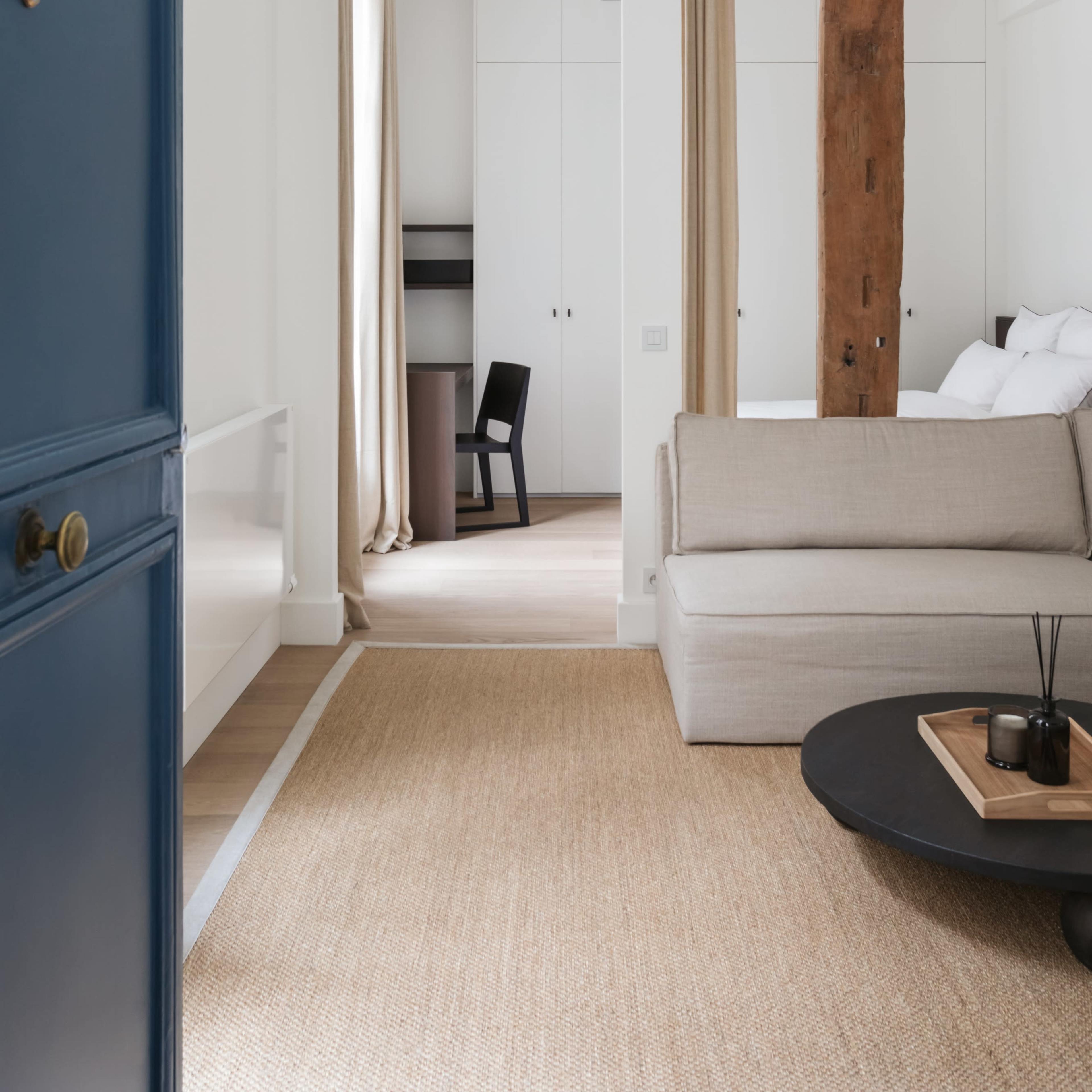 A blue door leads into a minimalist room featuring a sofa, coffee table, and a view of a study area with a chair and light-colored curtains.