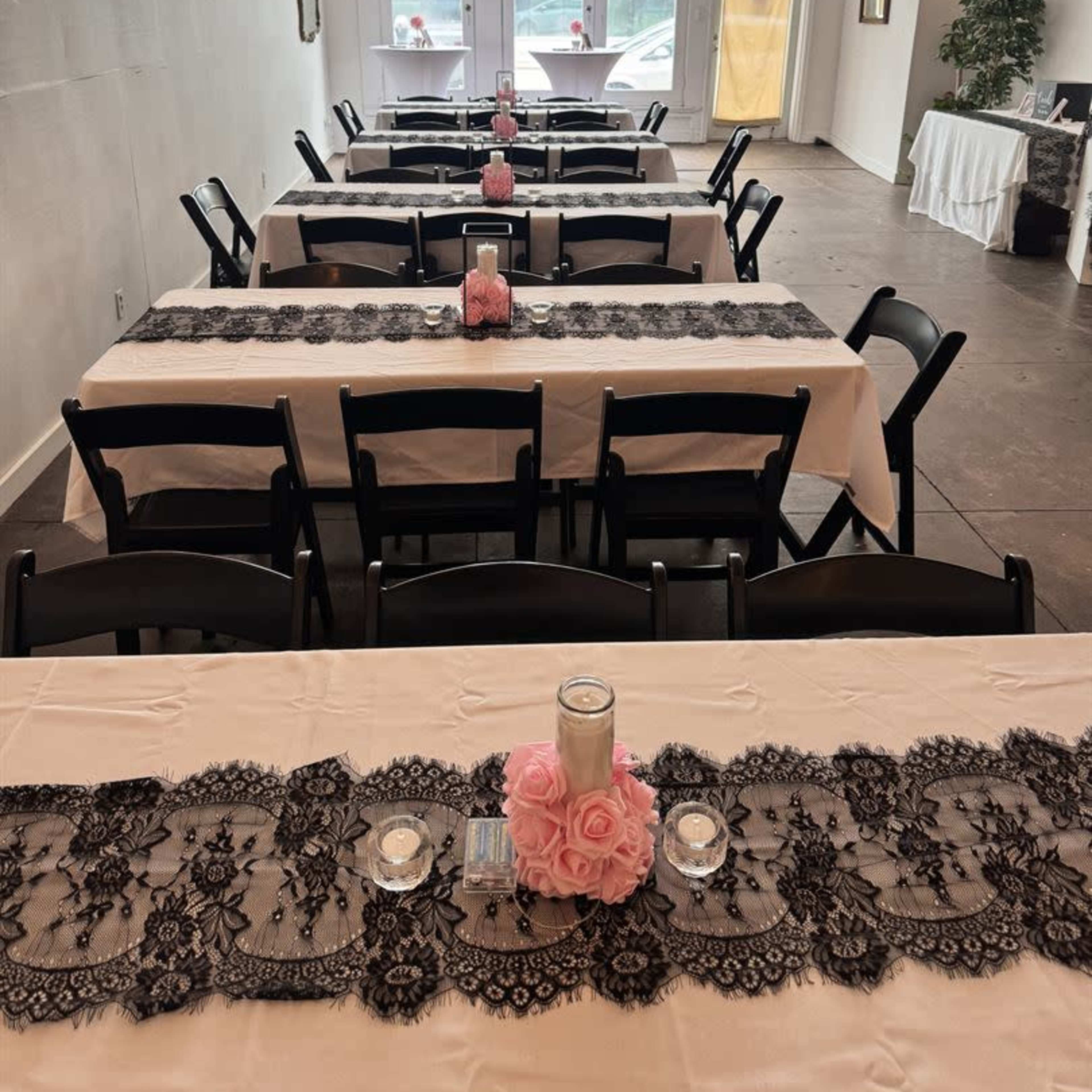 A decorative banquet room features several long tables set with lace runners, candles, and pink roses.