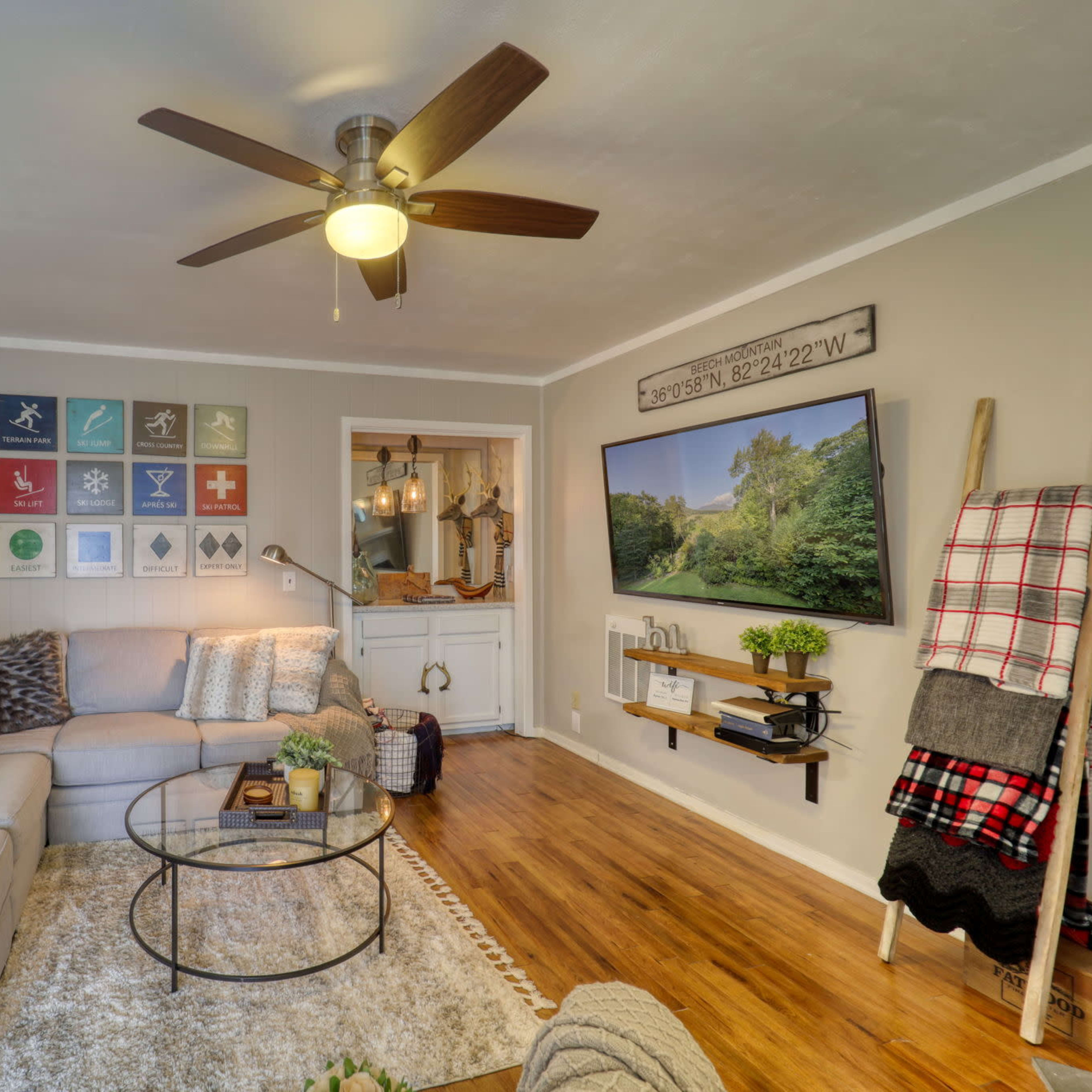 The living room features a light-colored sectional sofa, a round coffee table, wall art organized in a grid, and a flat-screen TV mounted above a wooden shelf.