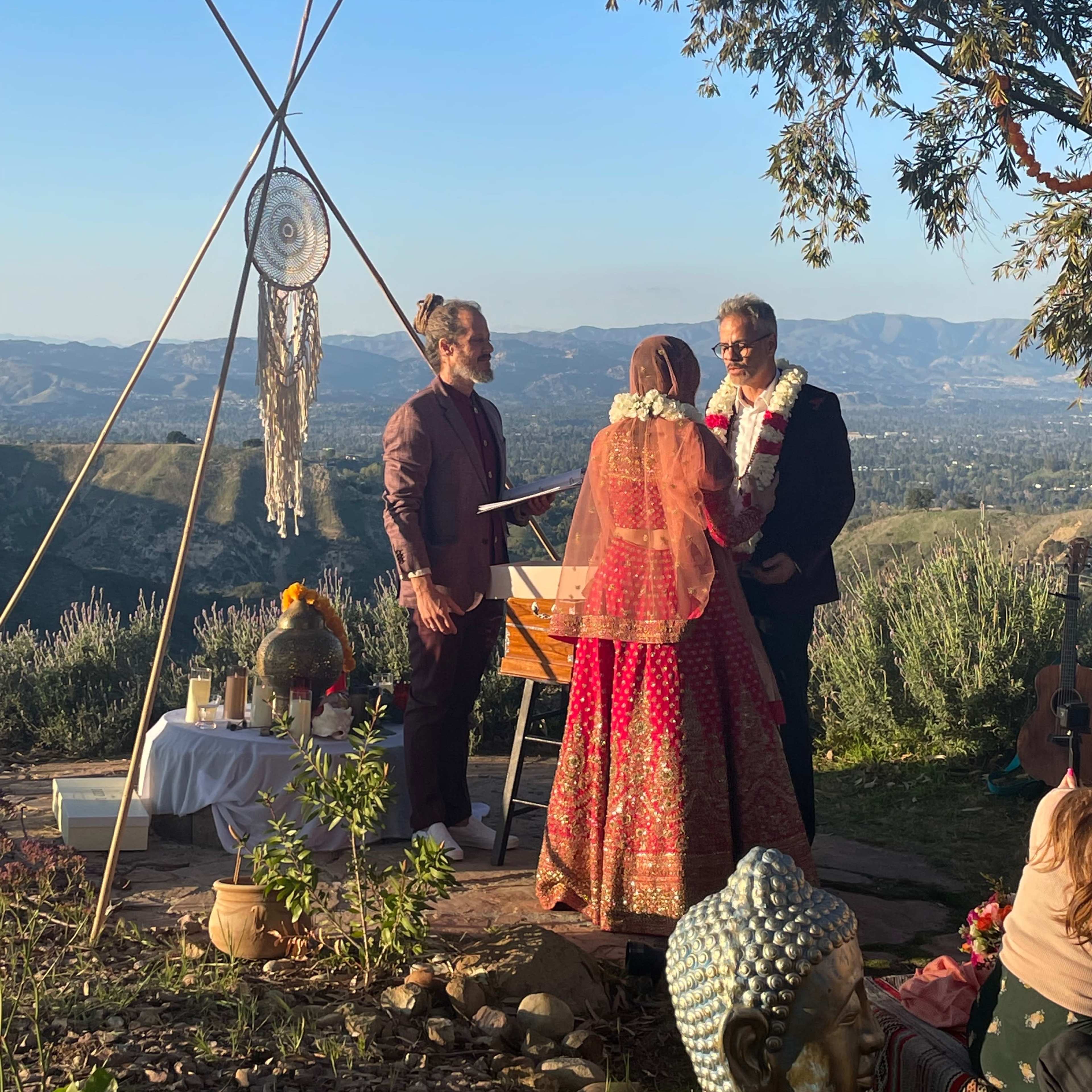 A wedding ceremony is taking place outdoors on a hillside with mountains in the background, featuring a couple standing at an altar adorned with decorative elements.