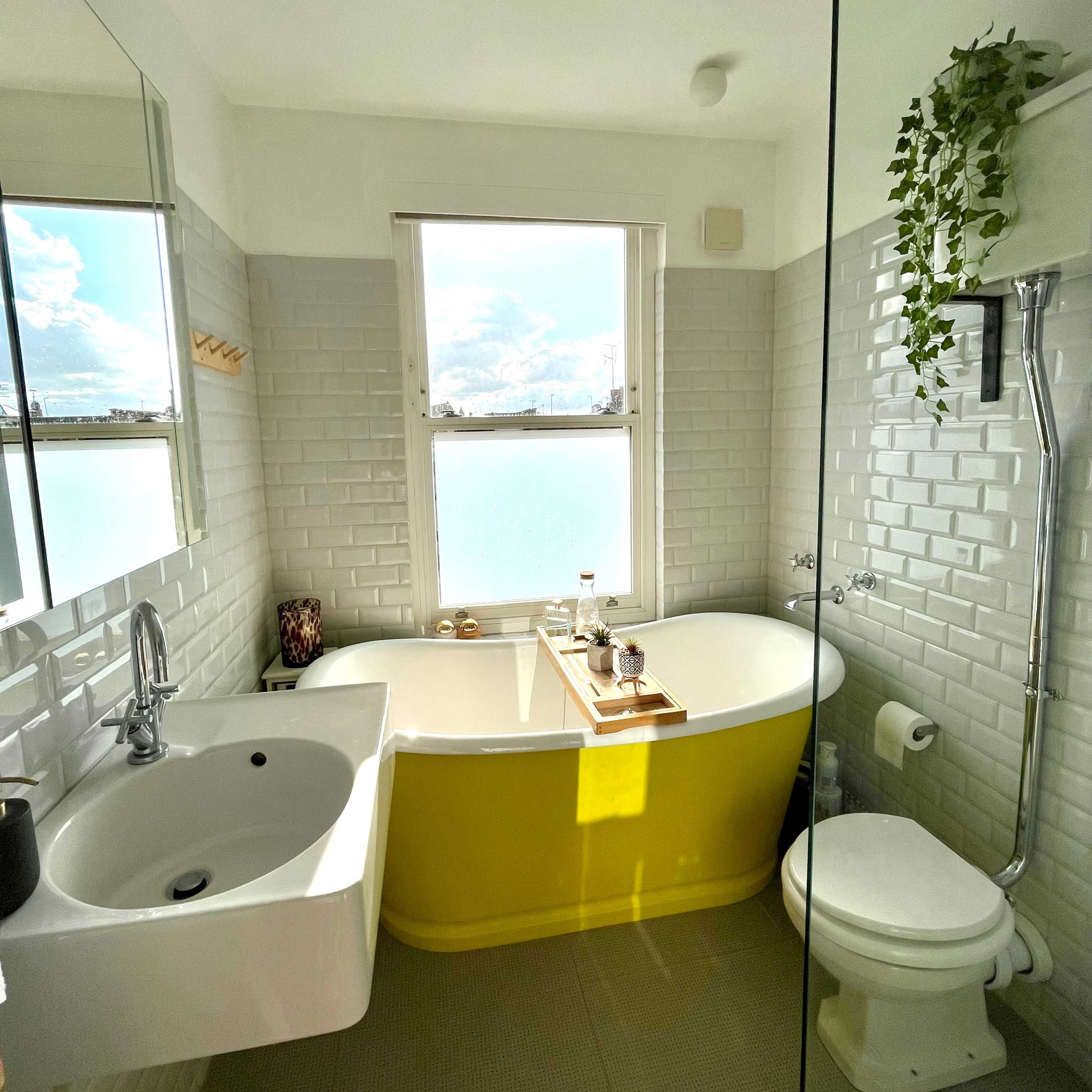 The bathroom features a freestanding yellow tub, a wall-mounted sink, and a glass shower enclosure with a large window allowing natural light.