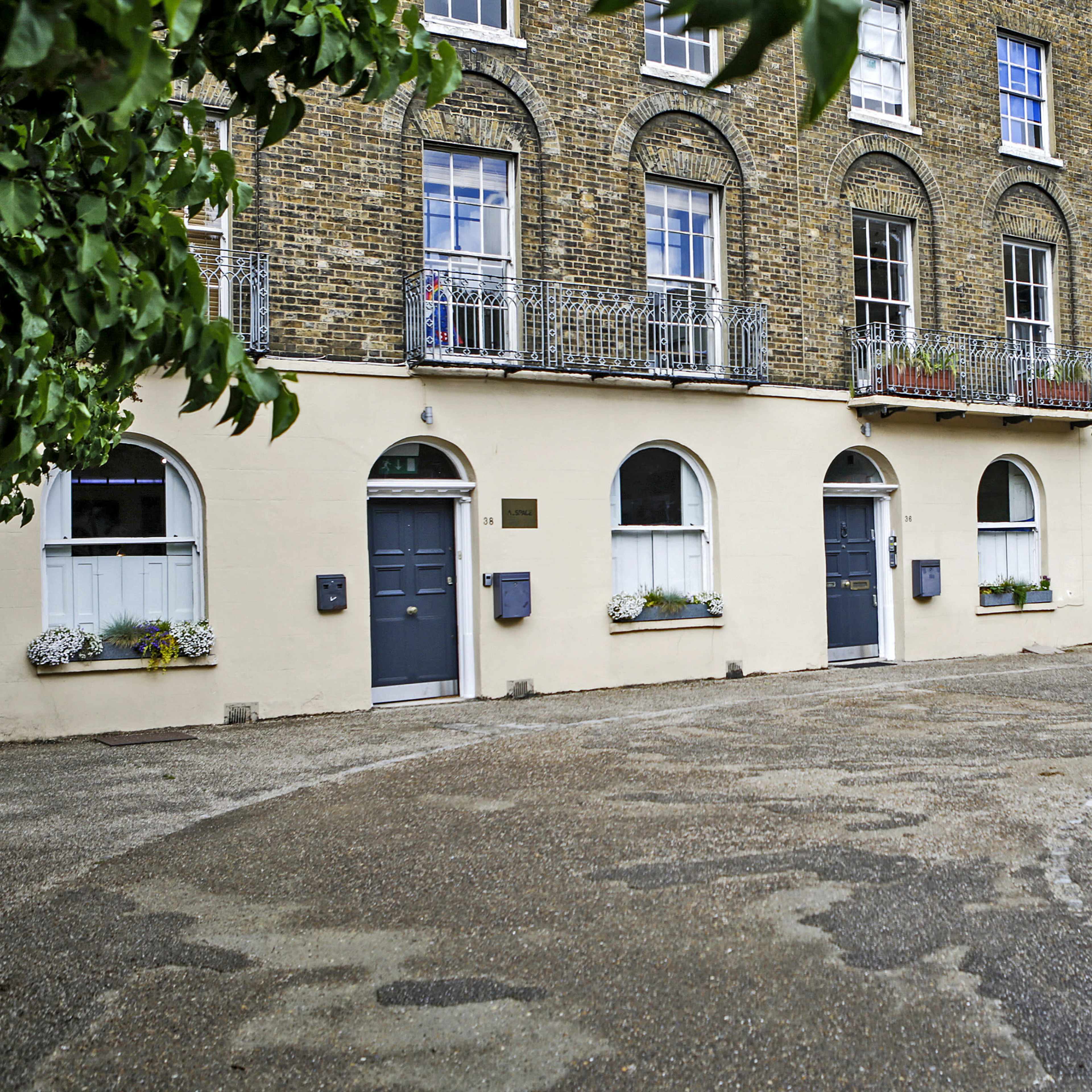 A row of four brownstone buildings with arched windows and blue doors, surrounded by a gravel path and greenery.