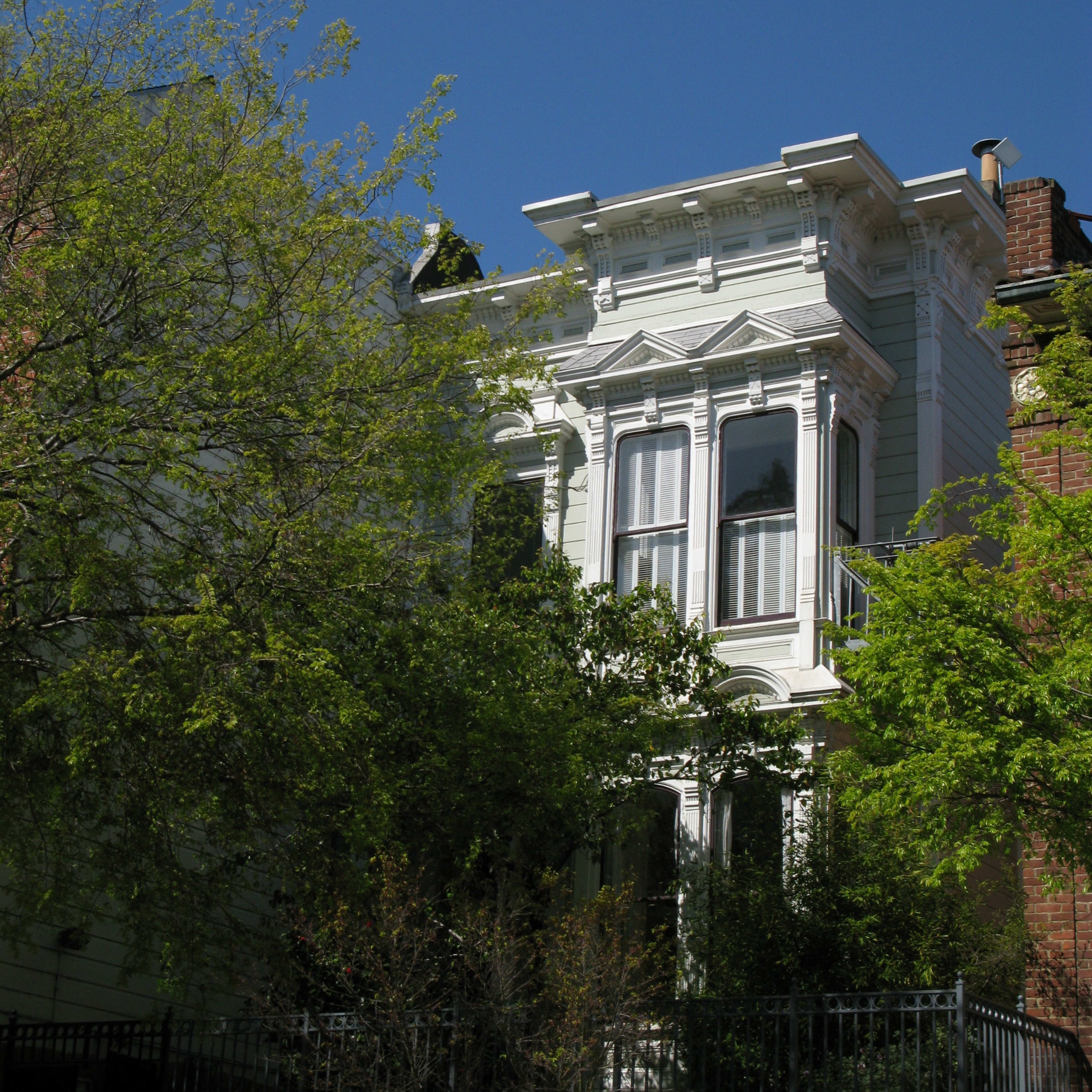 A white Victorian-style house is partially obscured by green foliage under a clear blue sky.