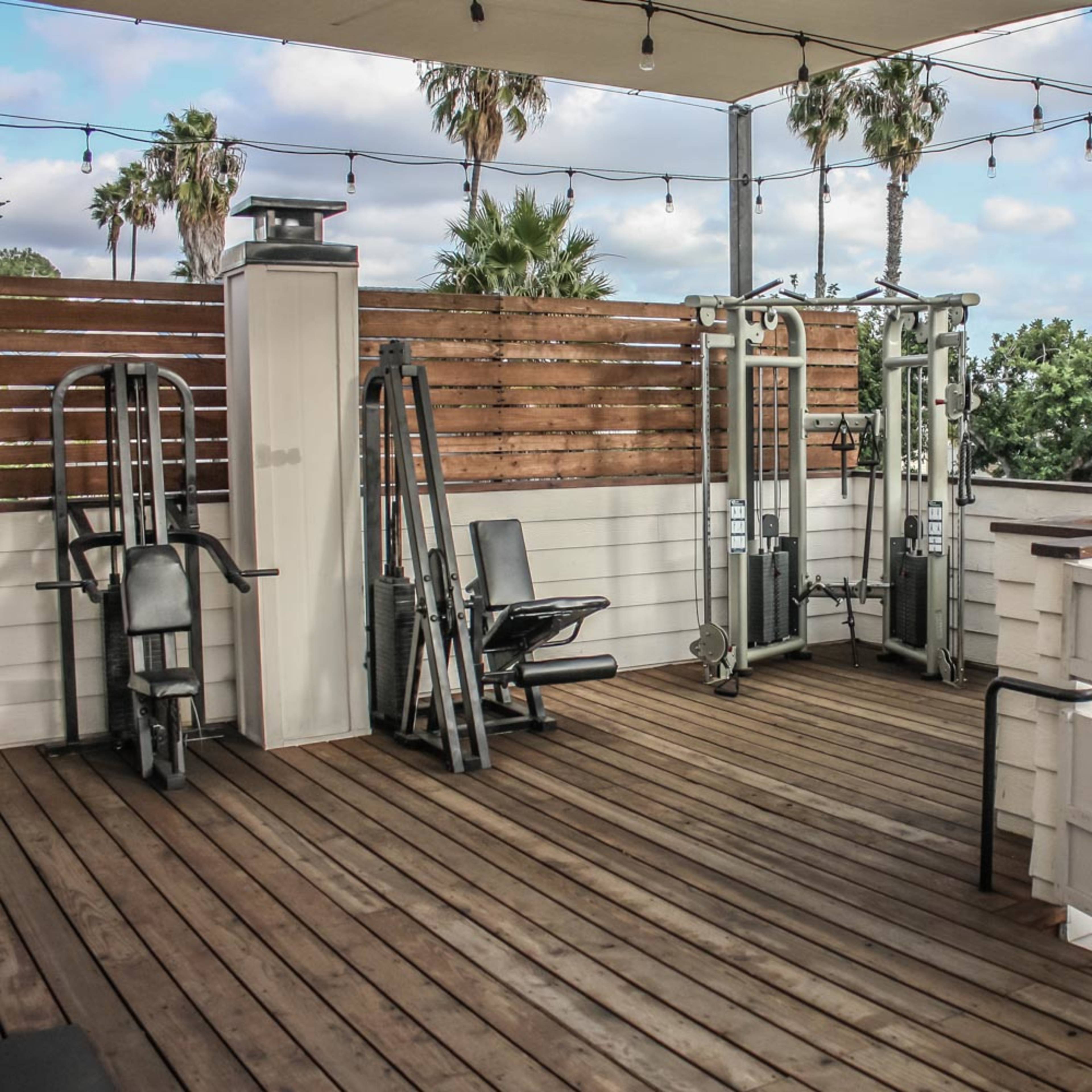 An outdoor gym area with various exercise equipment set on a wooden deck, surrounded by palm trees and string lights.