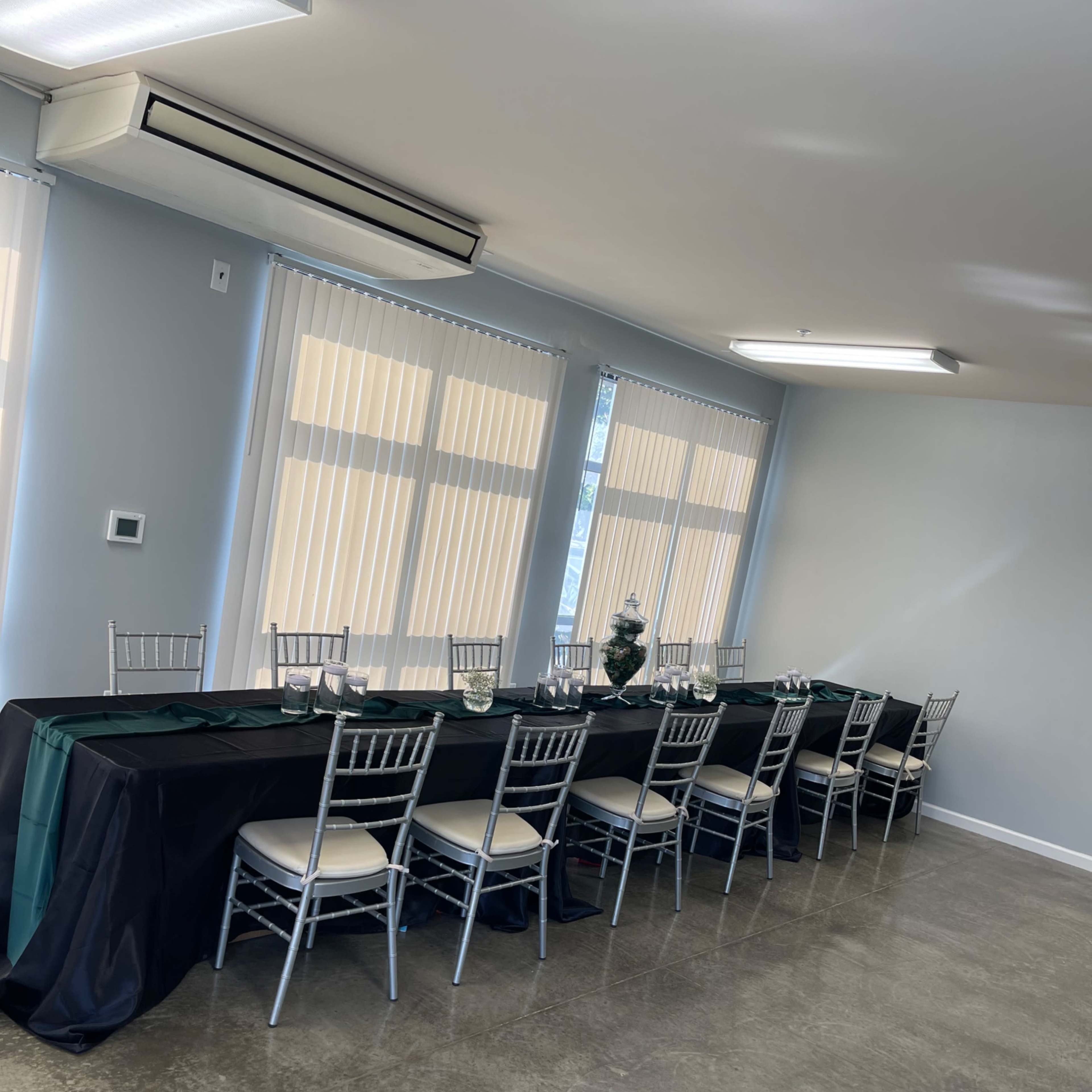 A long dining table with silver chairs is set against a wall of large windows in a well-lit room.