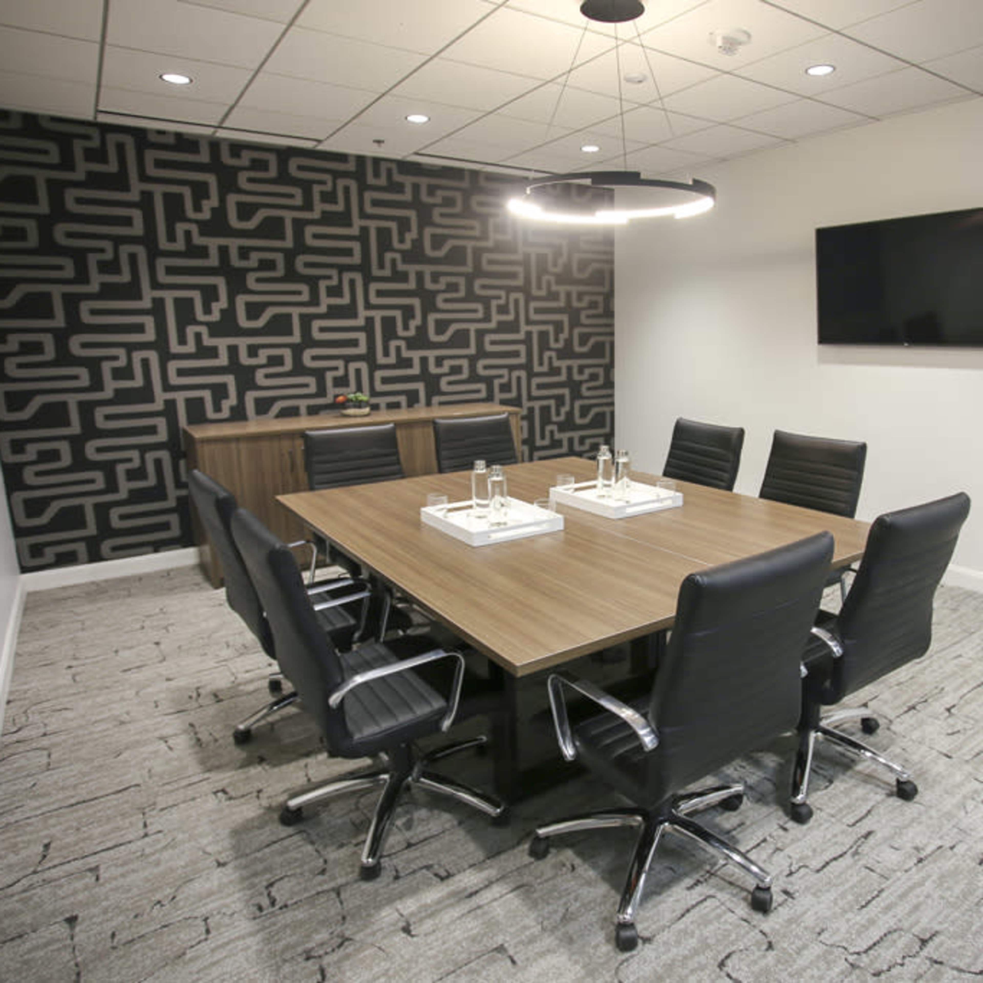 A modern conference room with a large rectangular wooden table surrounded by black office chairs, a patterned accent wall, and a flat-screen TV mounted on the wall.