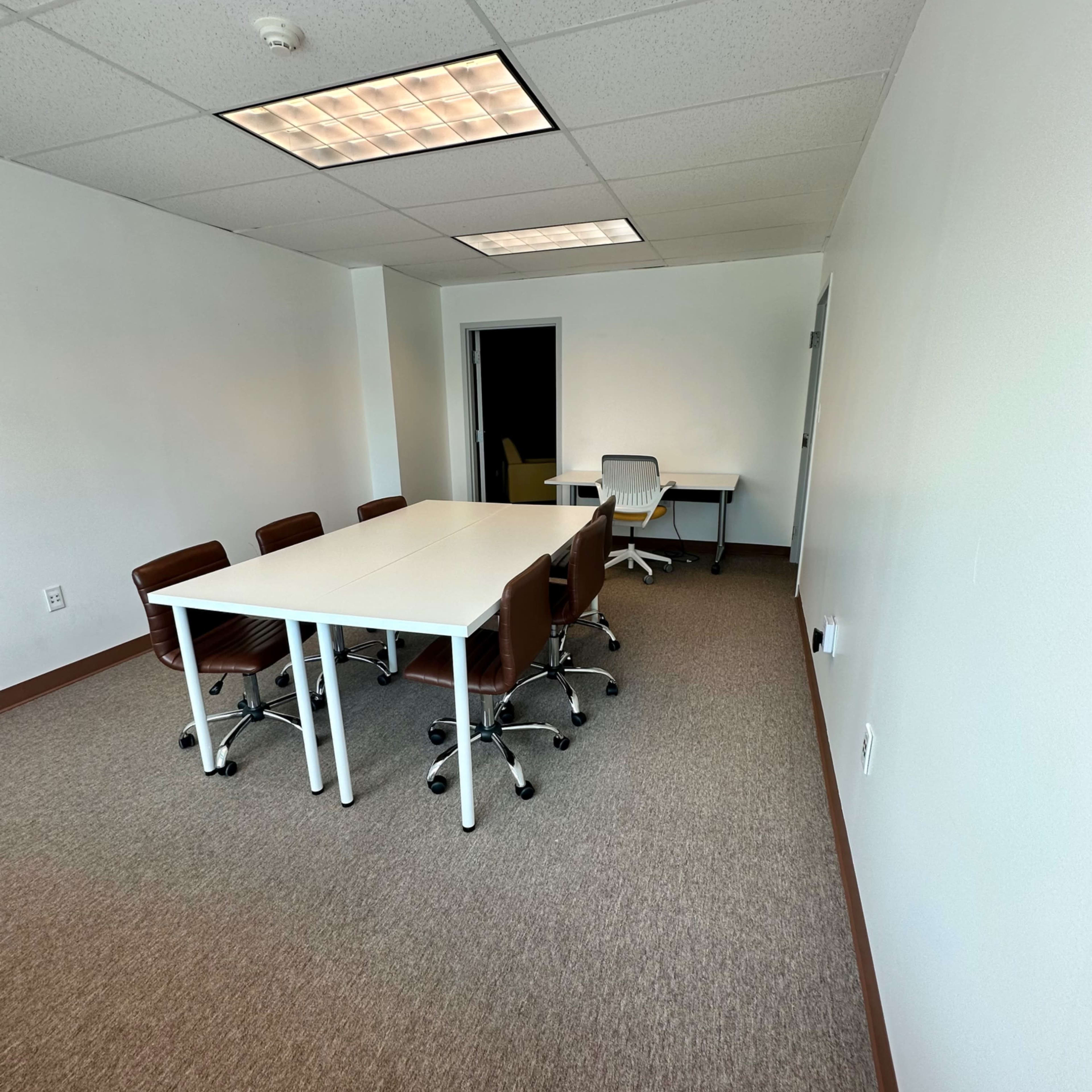A conference room with a white table surrounded by brown chairs and an additional desk in the corner.