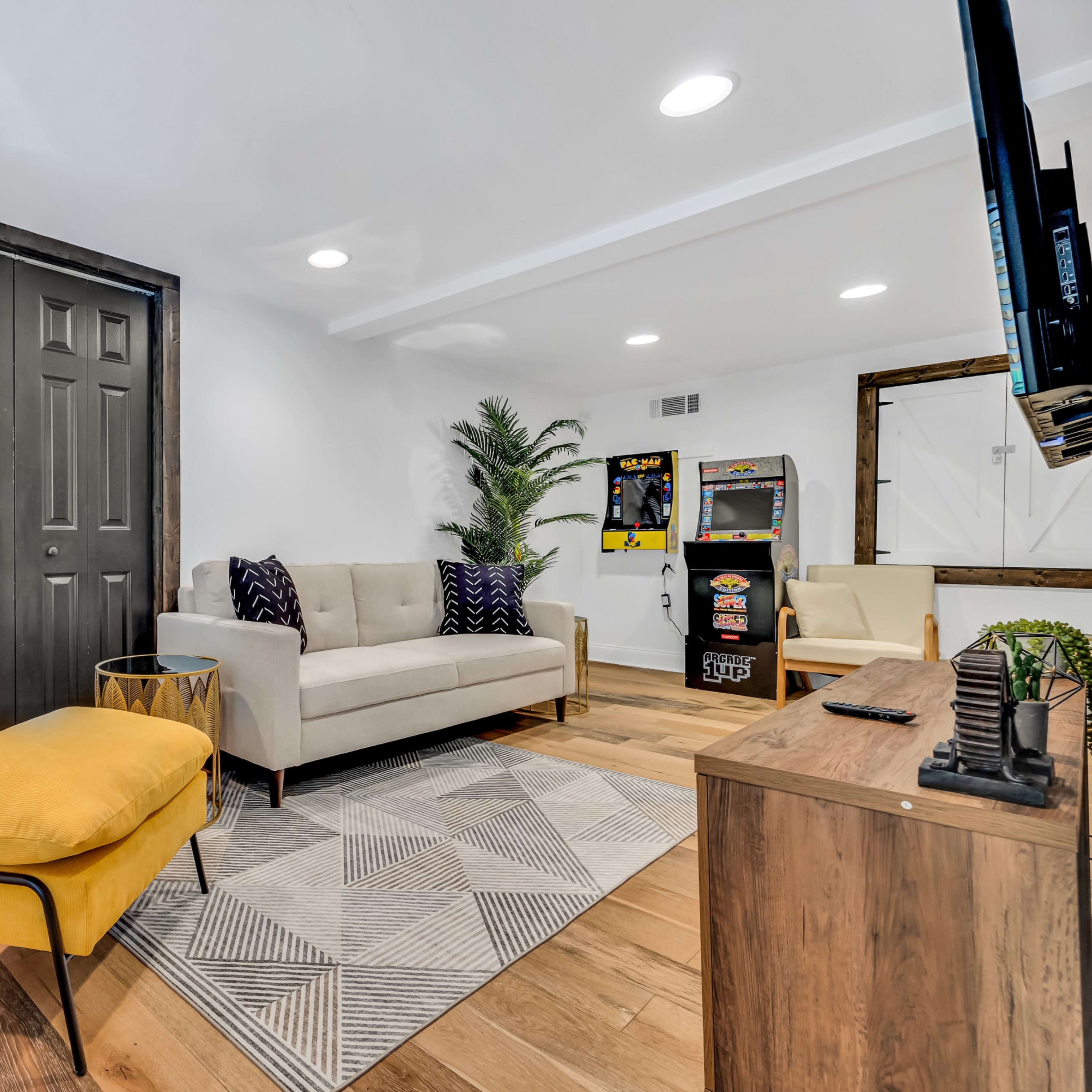 A cozy living room with a yellow armchair, a beige sofa, a patterned rug, and an arcade game machine in the corner.