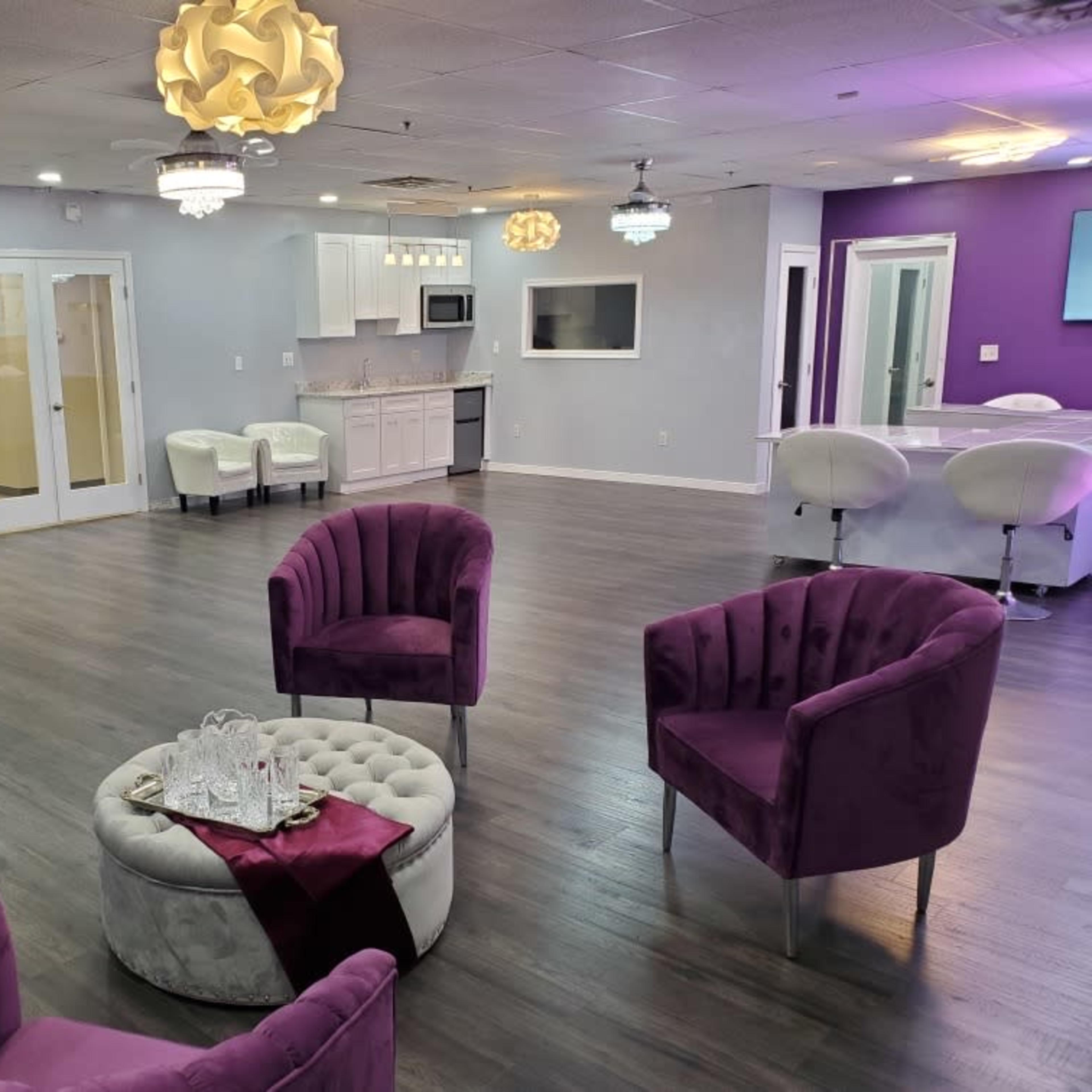 A stylish room with purple accent chairs, a circular ottoman, and a kitchenette, illuminated by decorative light fixtures.