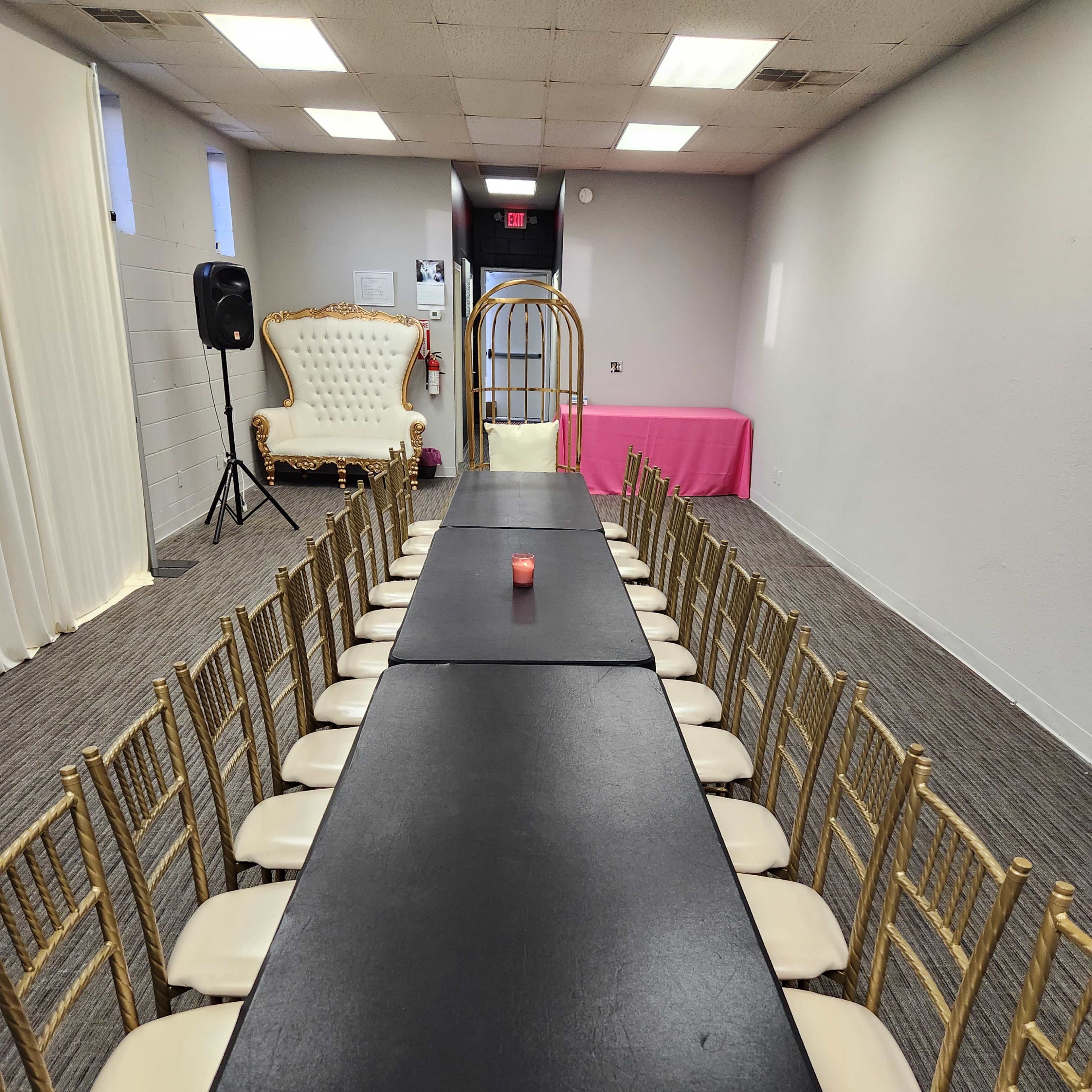 A long black table surrounded by gold chairs is set in a room with a white throne-style chair in the corner and a pink table at the far end.