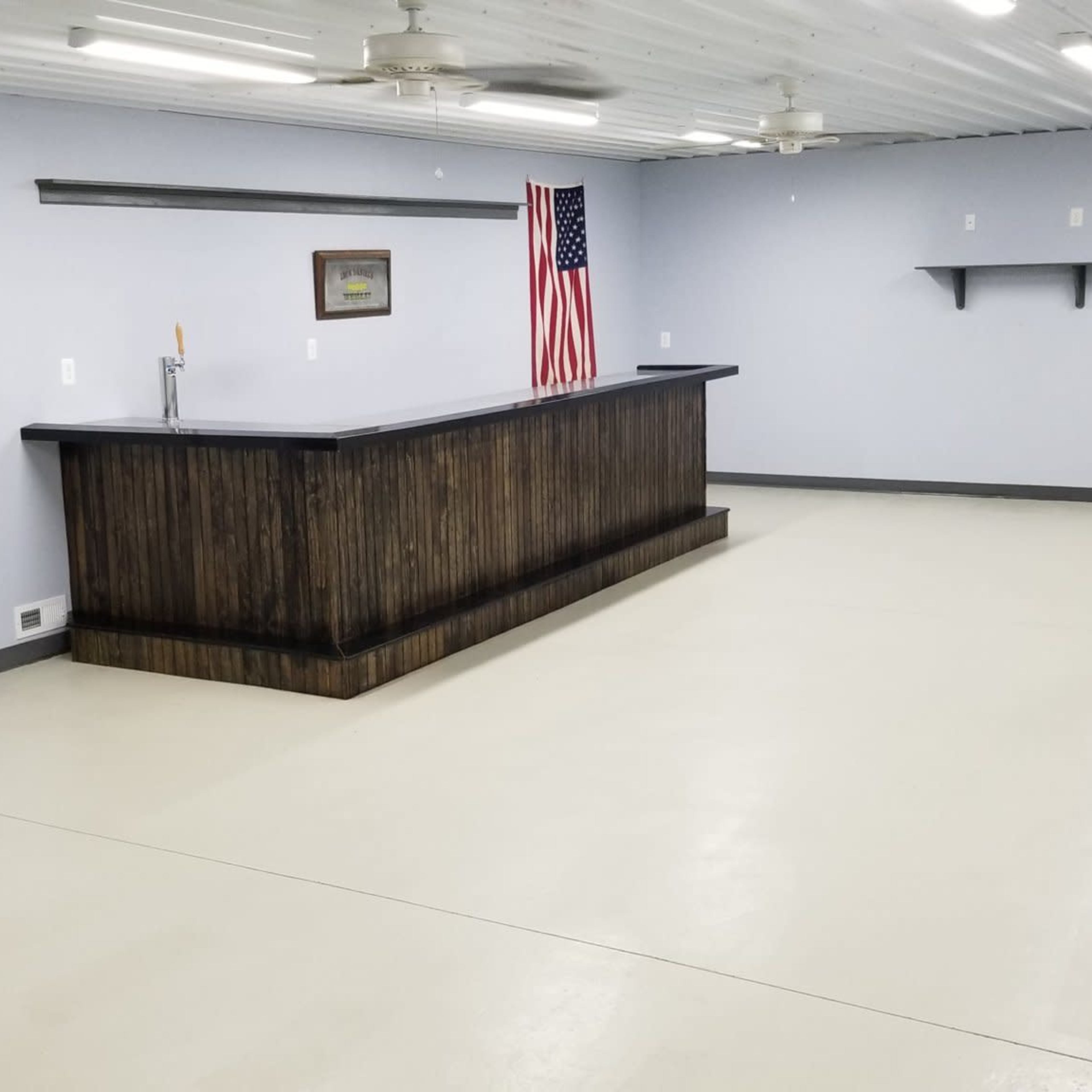 An empty room with a wooden bar at one end, a shelf on the wall, and an American flag displayed nearby.