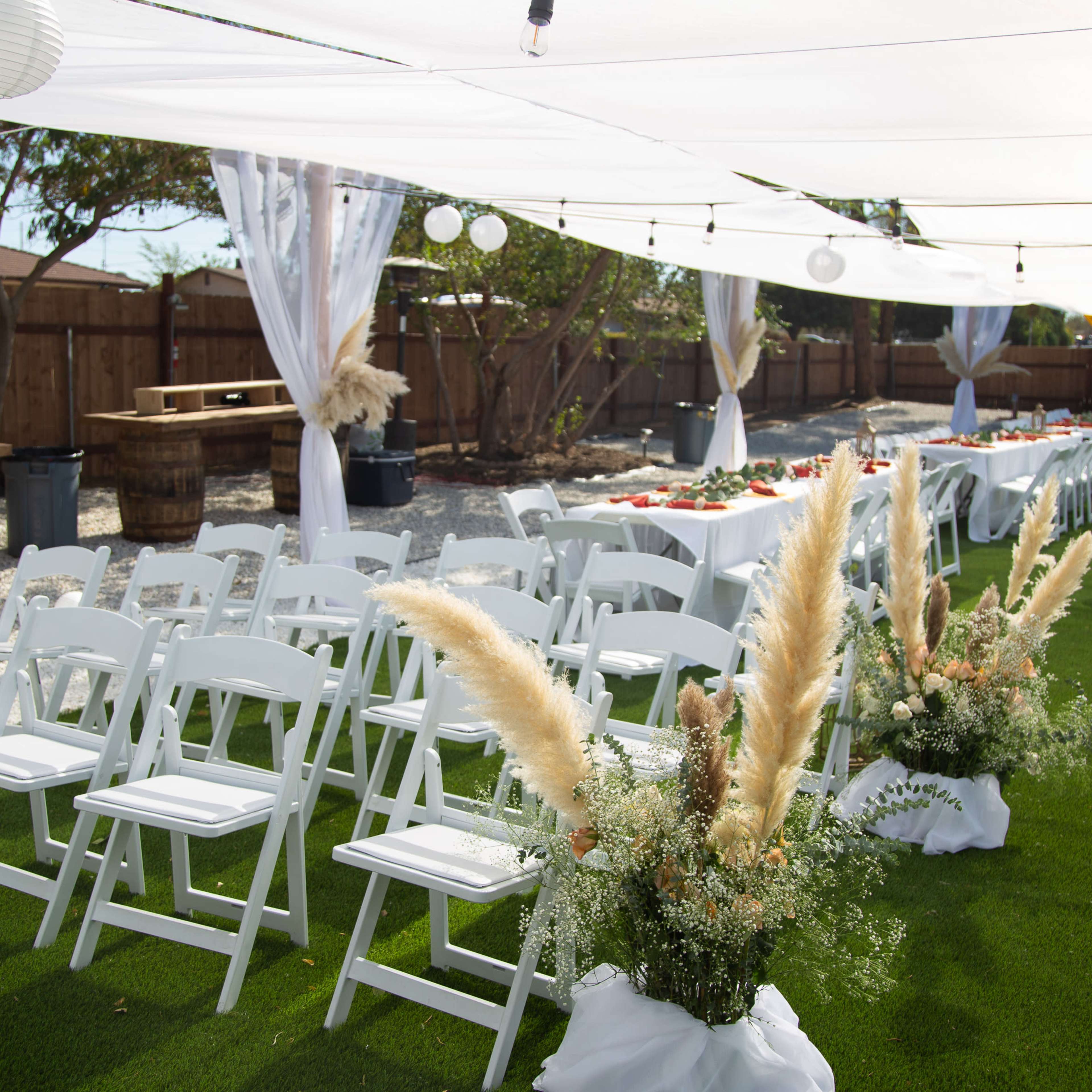 A decorated outdoor space features white folding chairs arranged for a gathering, surrounded by greenery and adorned with decorative elements including pampas grass and hanging lights.