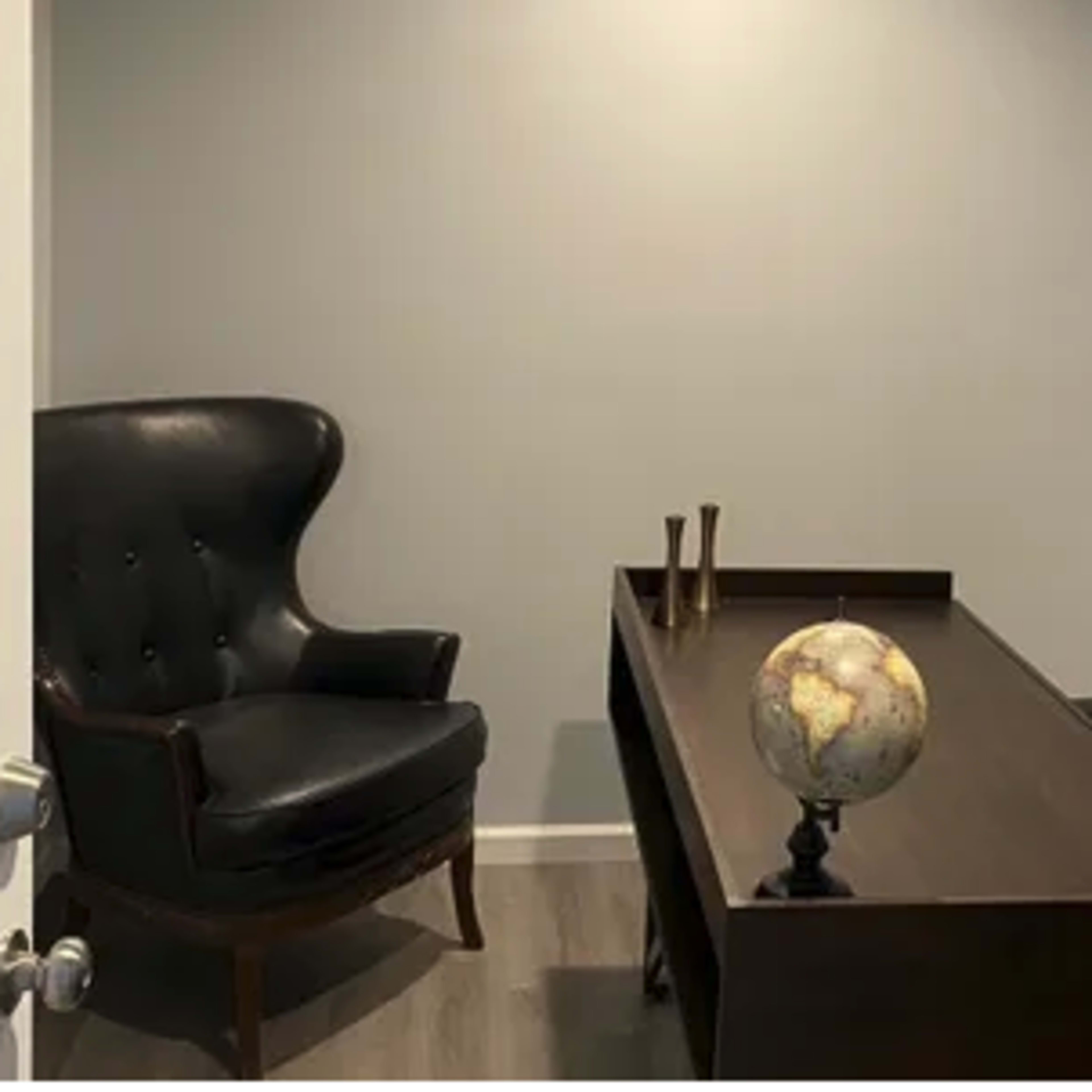 A small office space featuring a dark wooden desk, a globe on a stand, a black armchair, and a black office chair against a light gray wall.