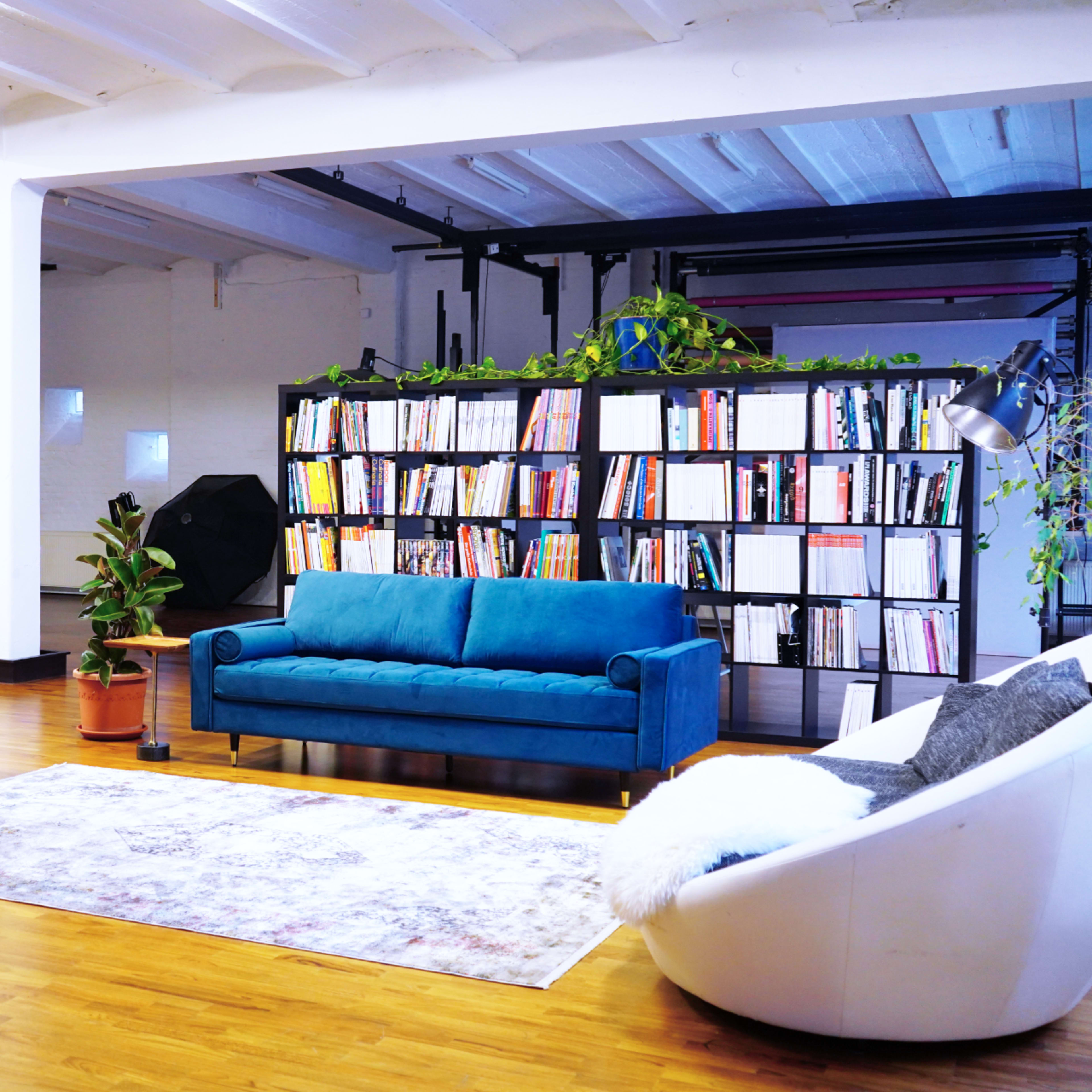 A spacious interior with a blue sofa, a modern bookshelf filled with books, various potted plants, and hardwood flooring.
