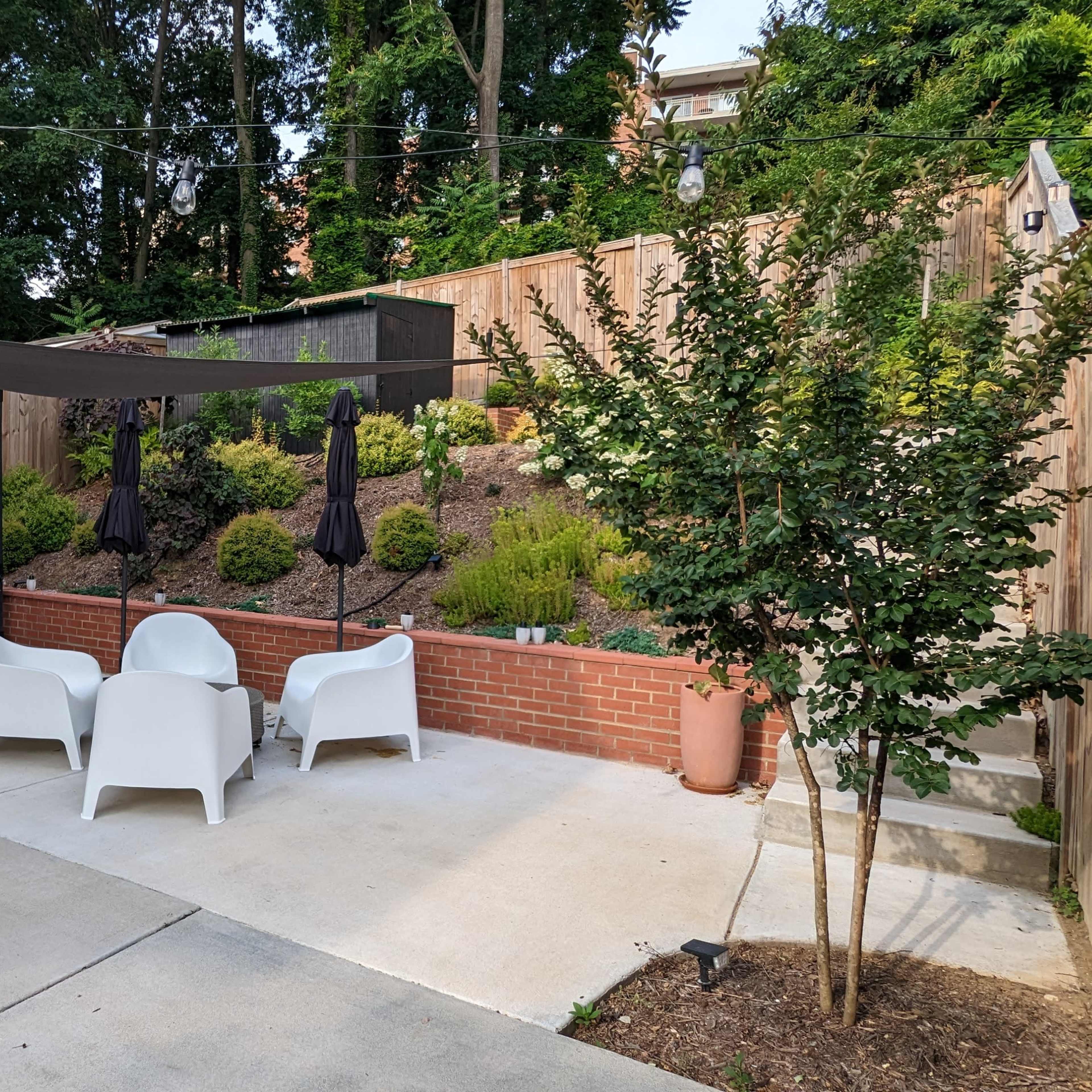A landscaped patio area featuring white chairs set around a small table, with a shaded canopy and decorative plants in the background.