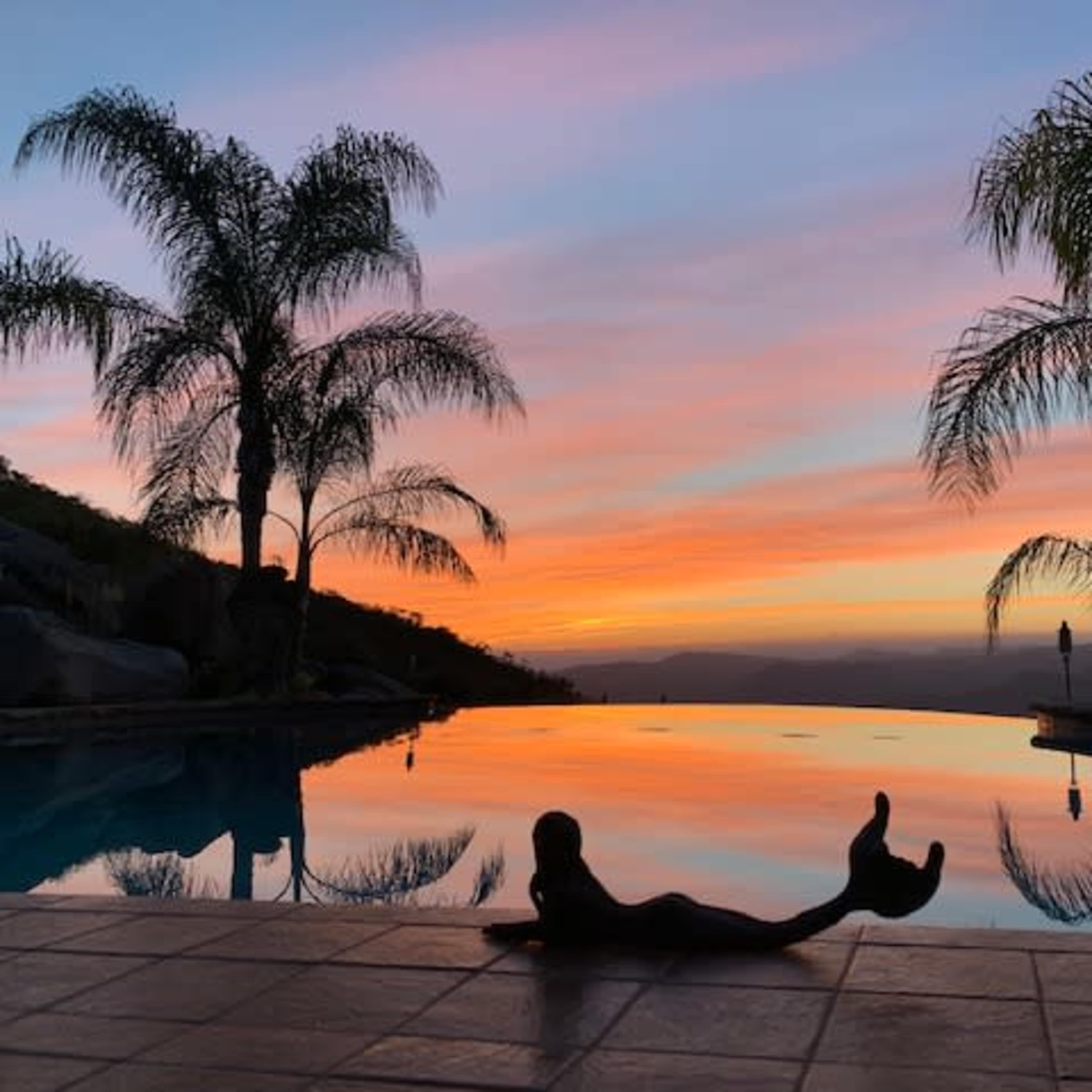 A silhouetted figure relaxes by a pool reflecting a sunset backdrop with palm trees on either side.