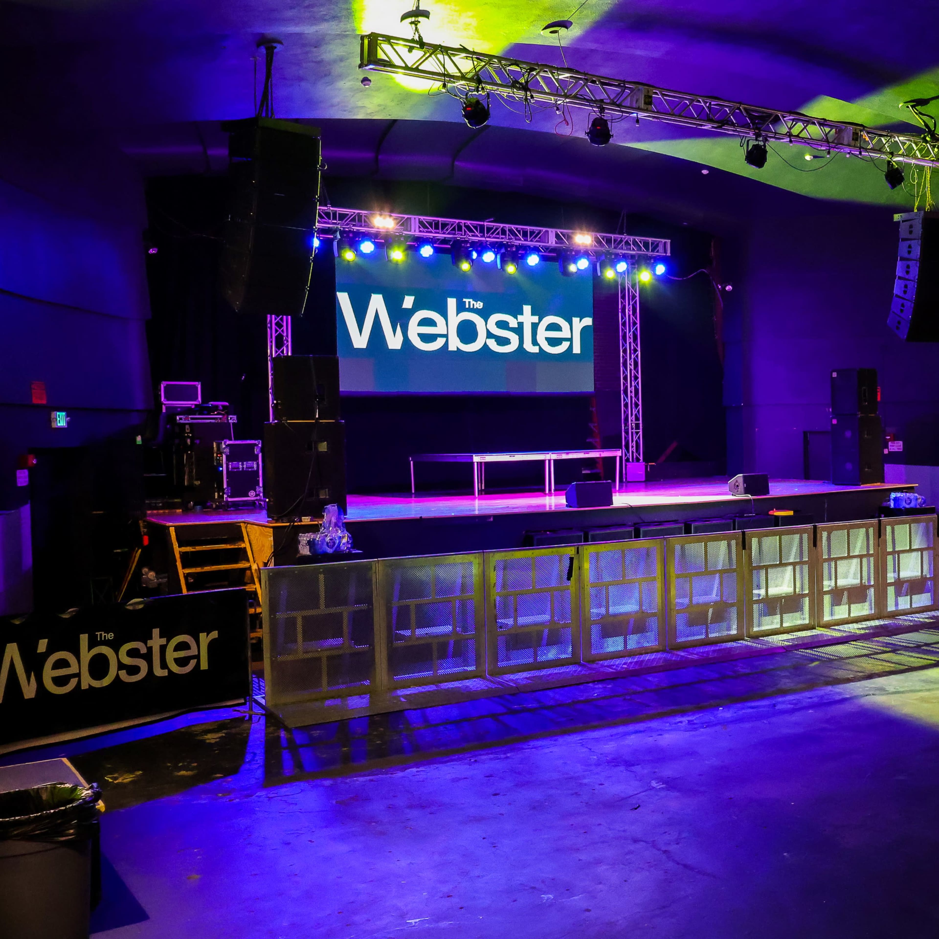 An empty concert stage at The Webster, with lighting set up and a large screen displaying the venue's name.
