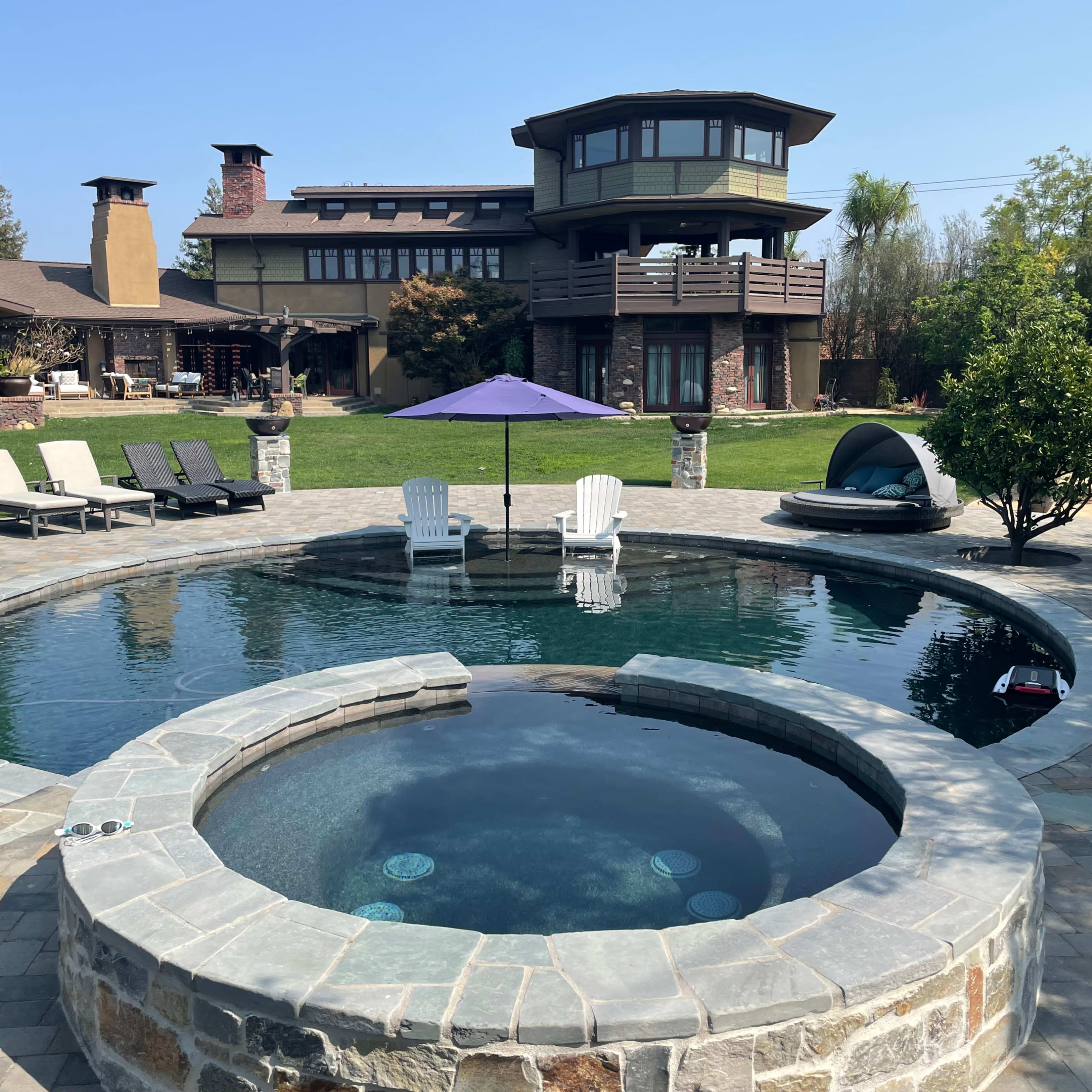 A circular pool with a spa area is surrounded by patio stones, lounge chairs, and a view of a multi-level house in the background.