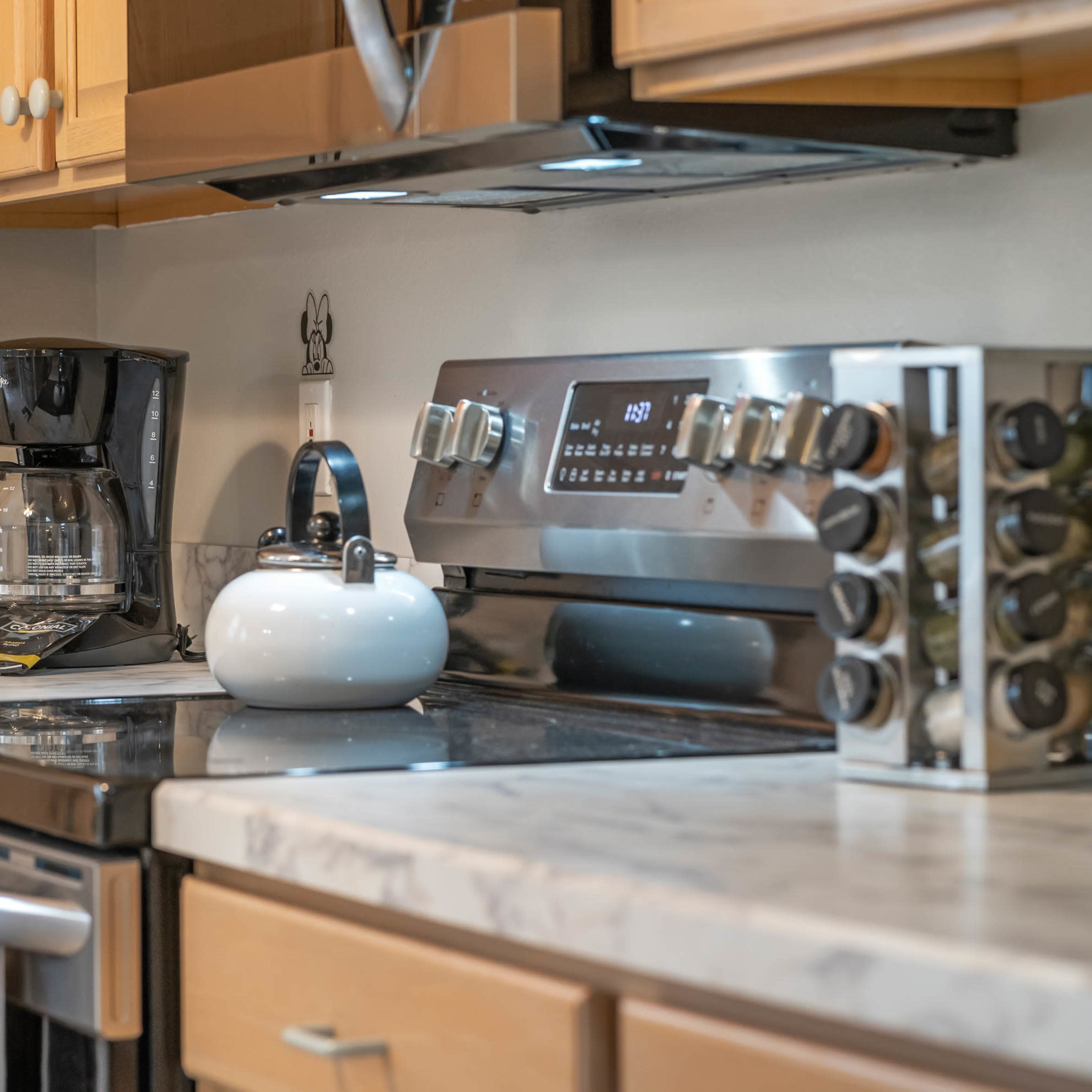 A kitchen countertop features a coffee maker, a kettle on the stove, and a variety of spice jars alongside a modern oven and cabinetry.