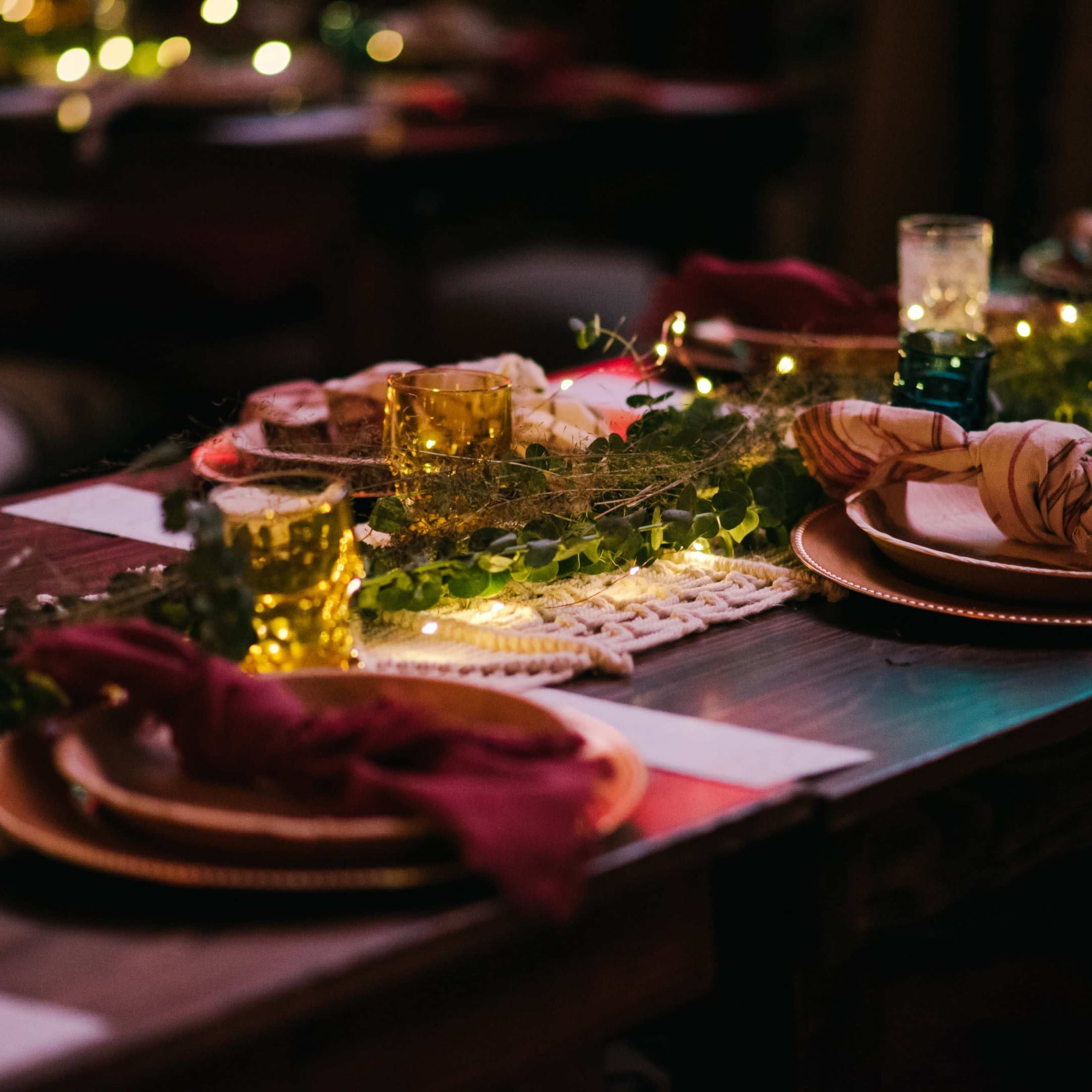 A long dining table is set with plates, candles, and green foliage, illuminated by string lights in a cozy setting.
