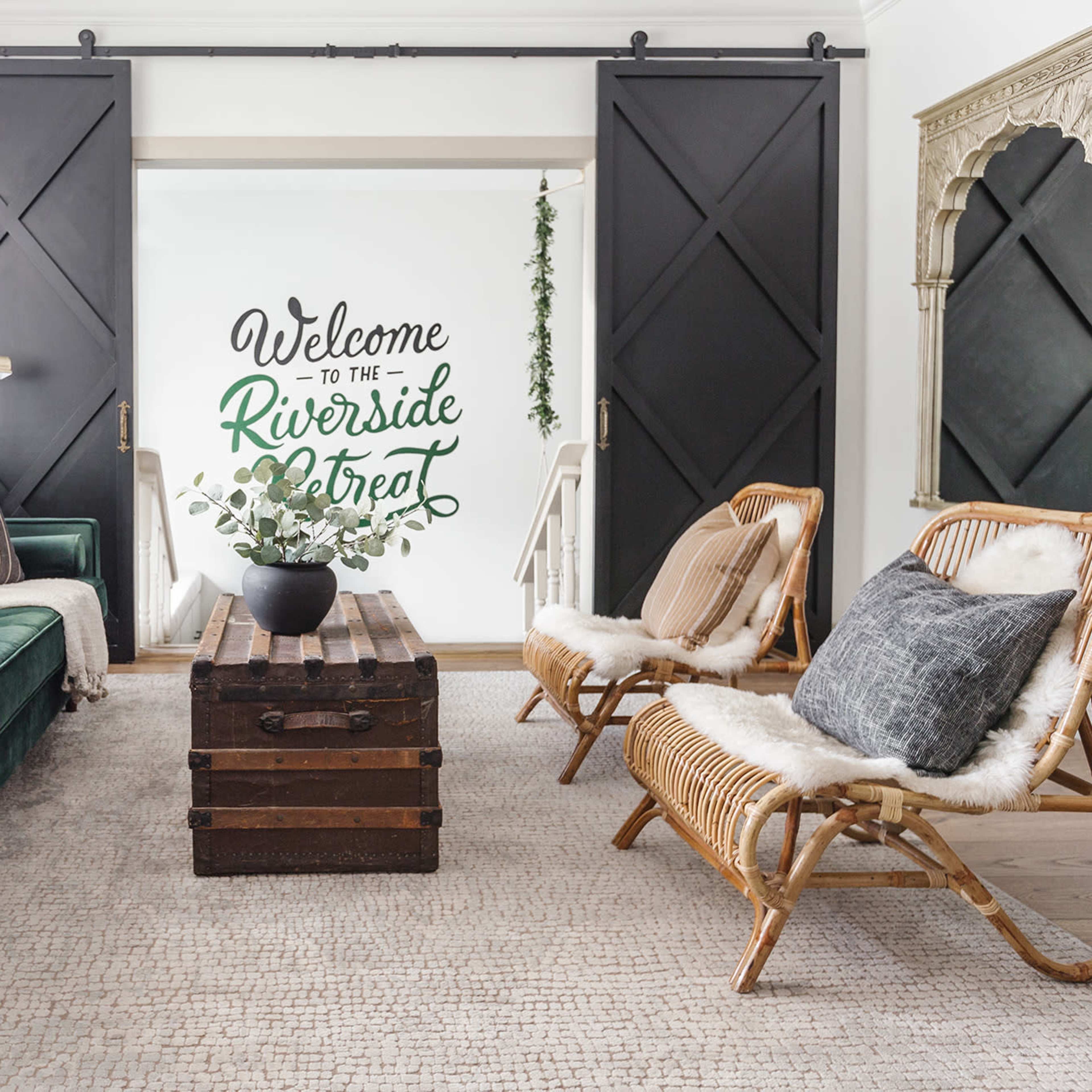 A living room with a green sofa, two rattan chairs, and a wooden coffee table, featuring a wall with the words "Welcome to the Riverside Street" painted on it.