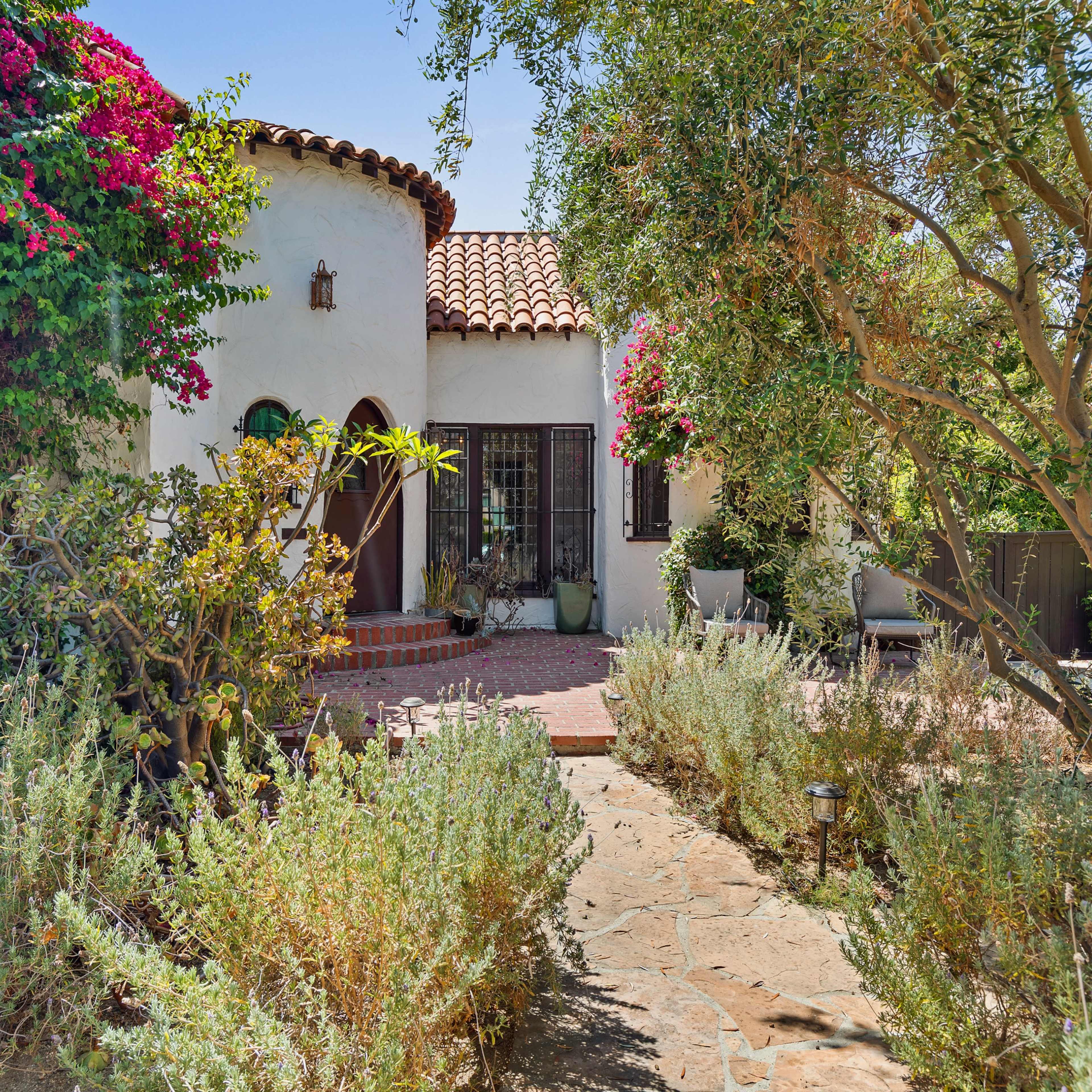 A Spanish-style house with a tiled roof is surrounded by blooming plants and a stone pathway.