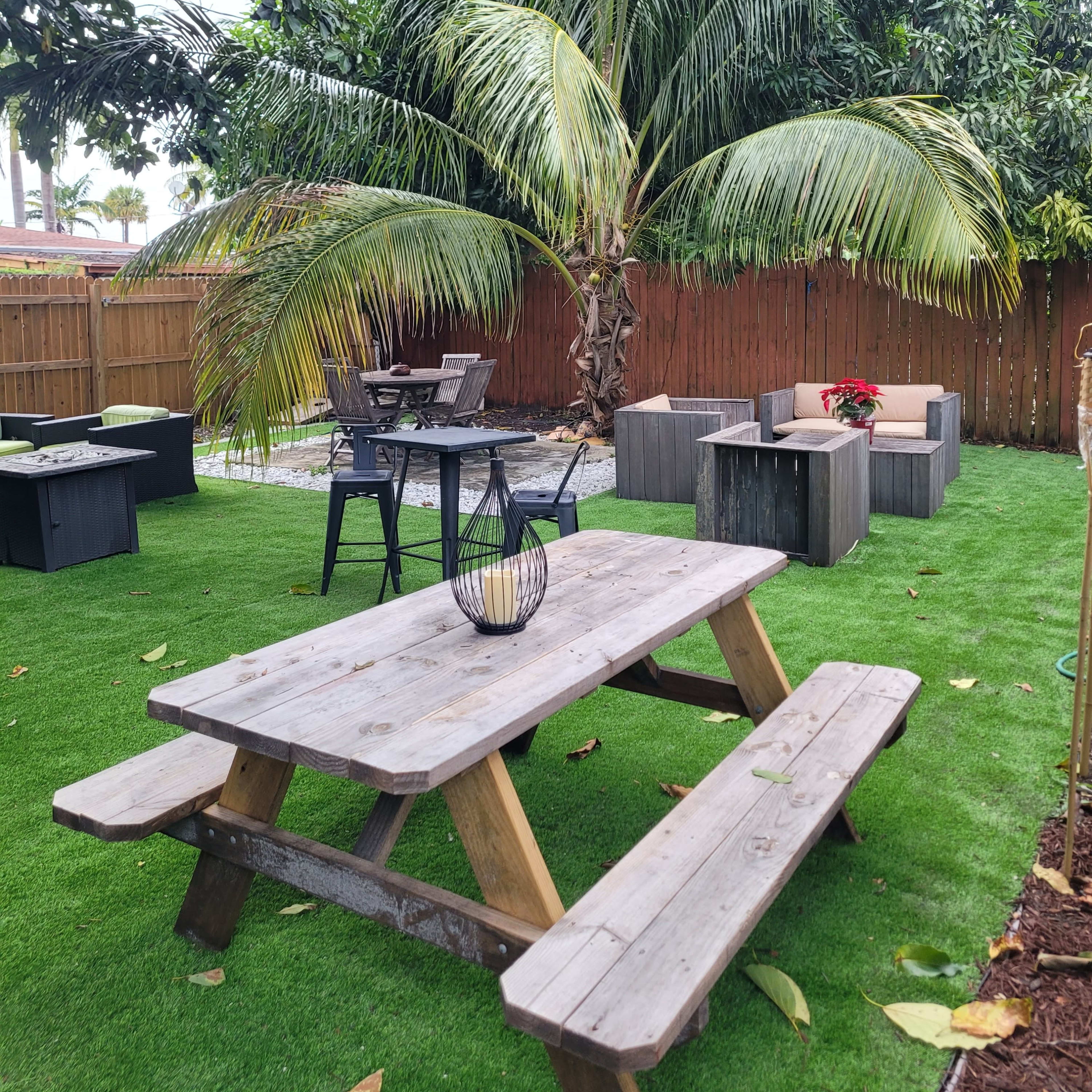 A wooden picnic table sits on a grassy area surrounded by palm trees and outdoor seating arrangements.