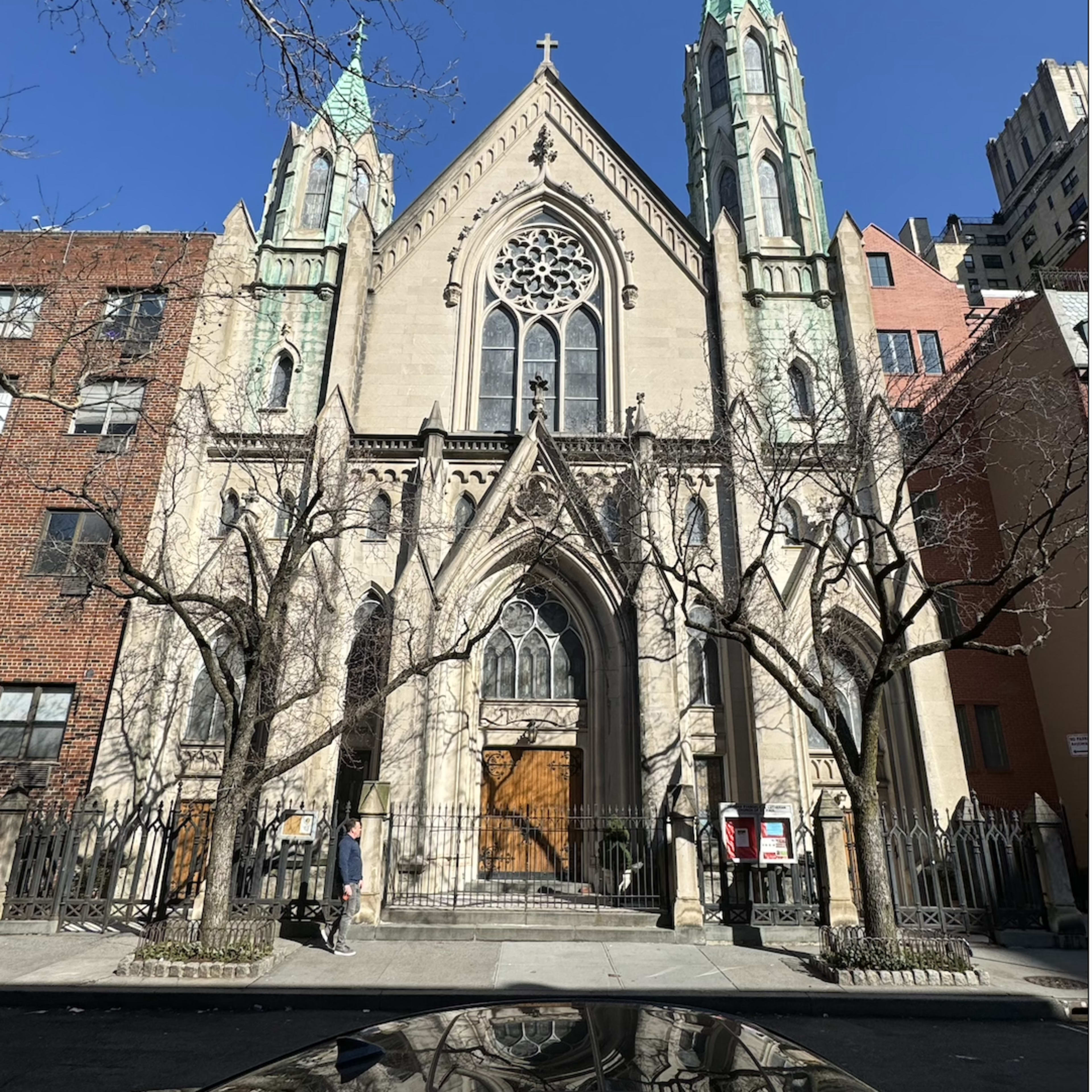 A Gothic-style church with pointed towers and intricate stained glass windows, located between urban buildings against a clear blue sky.