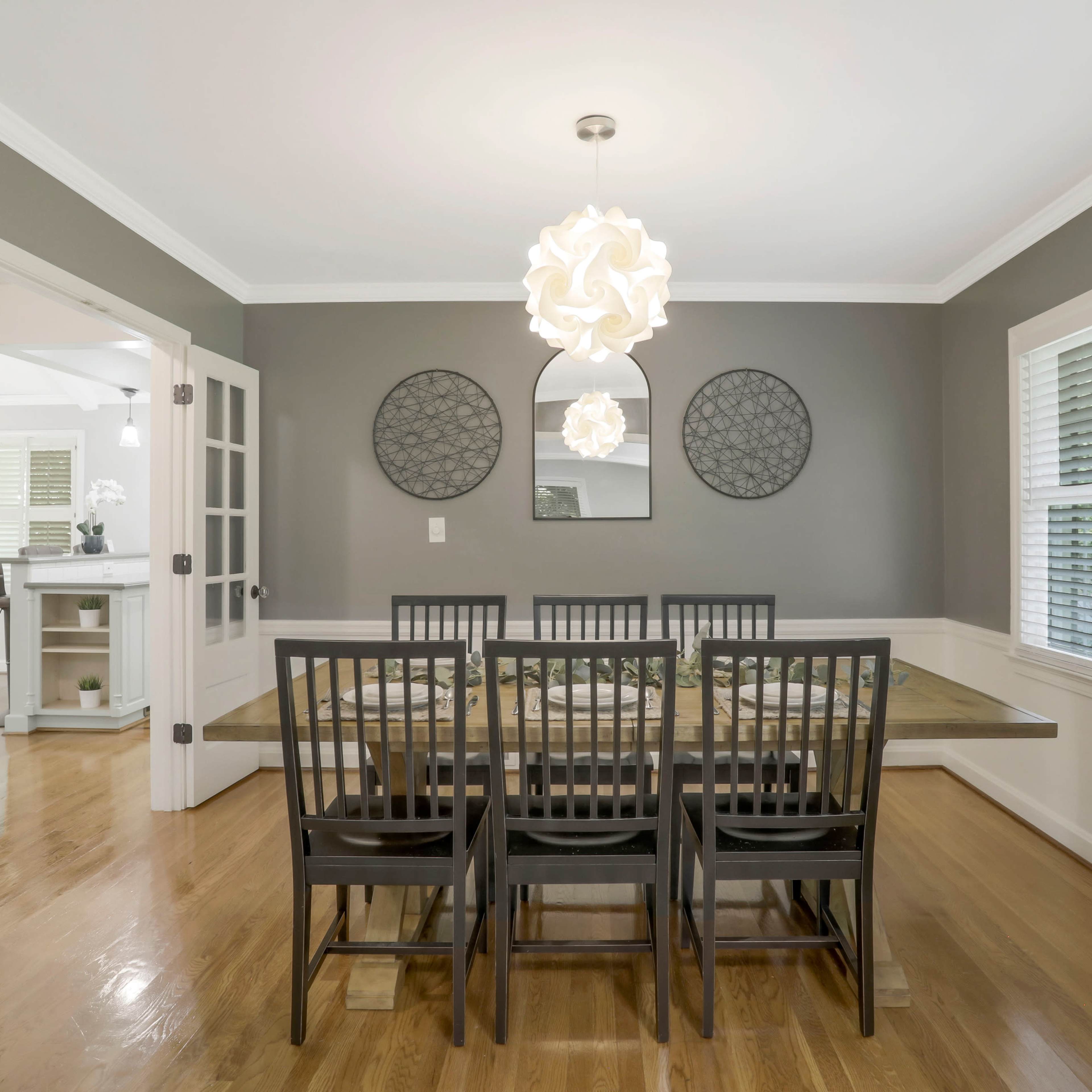 A dining room features a wooden table surrounded by six black chairs, with a modern light fixture overhead and decorative mirrors on the gray walls.