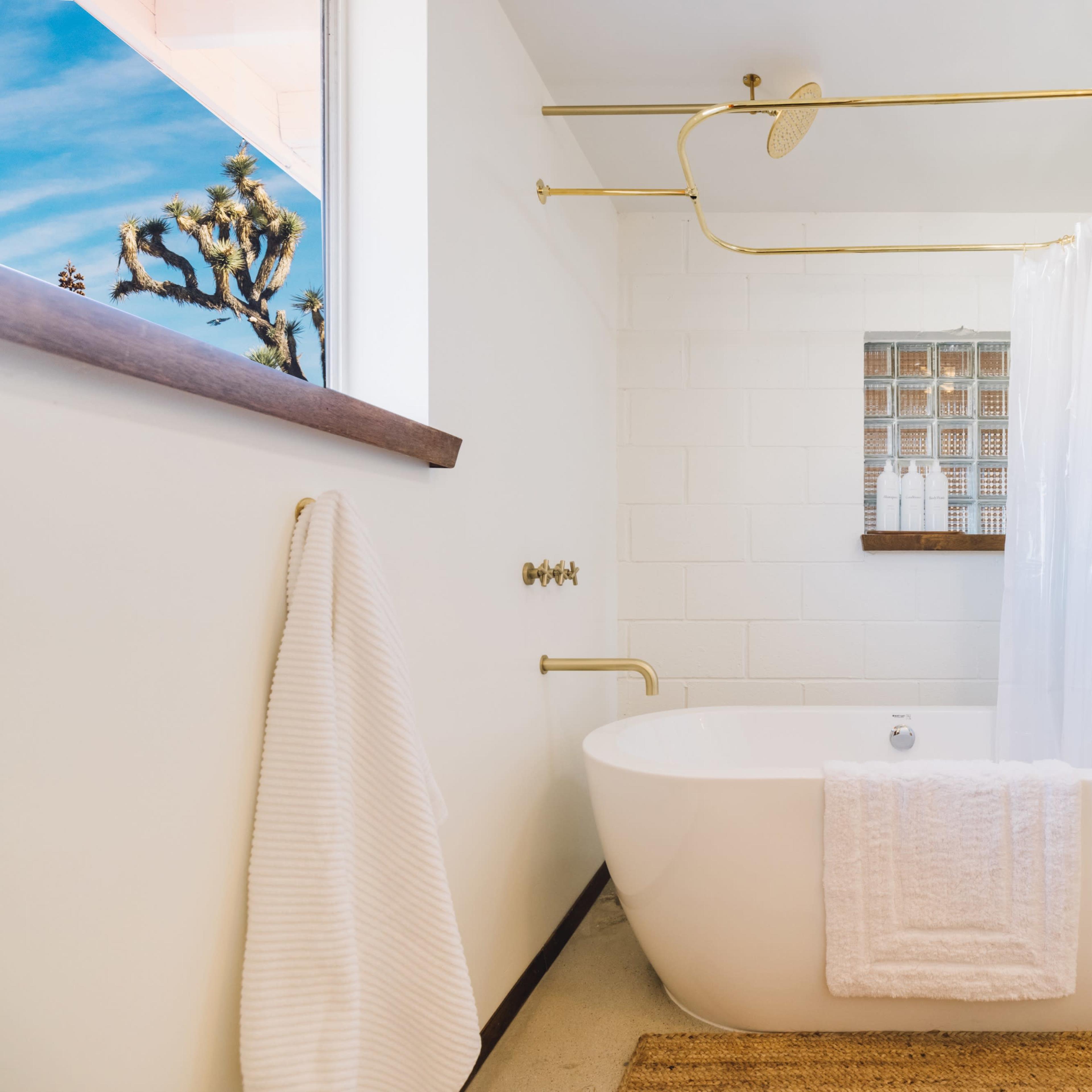 A modern bathroom features a freestanding white bathtub, a gold shower fixture, and a window with a view of a Joshua tree.
