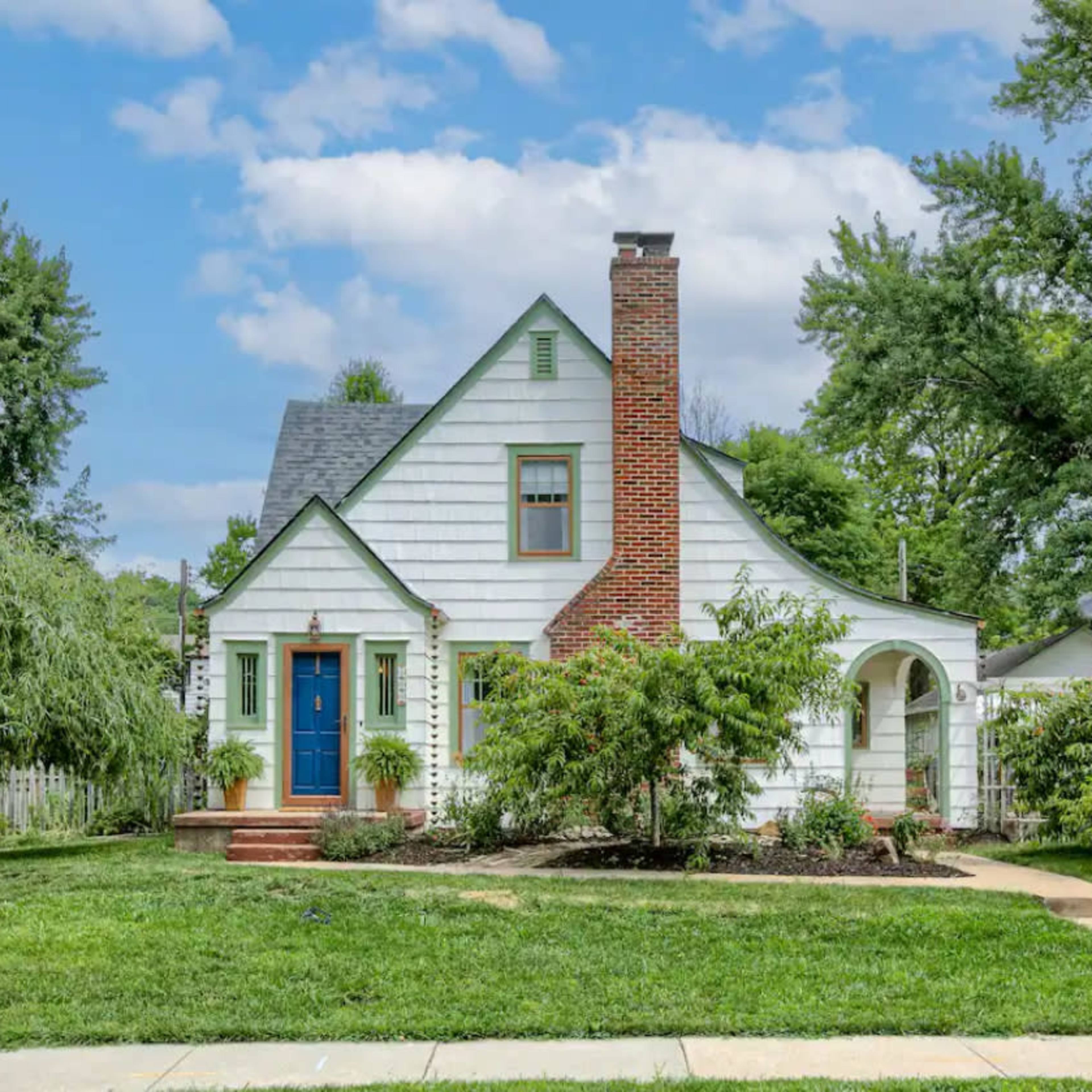A two-story, white clapboard house features a blue front door, a brick chimney, and a well-maintained front yard with trees and shrubs.