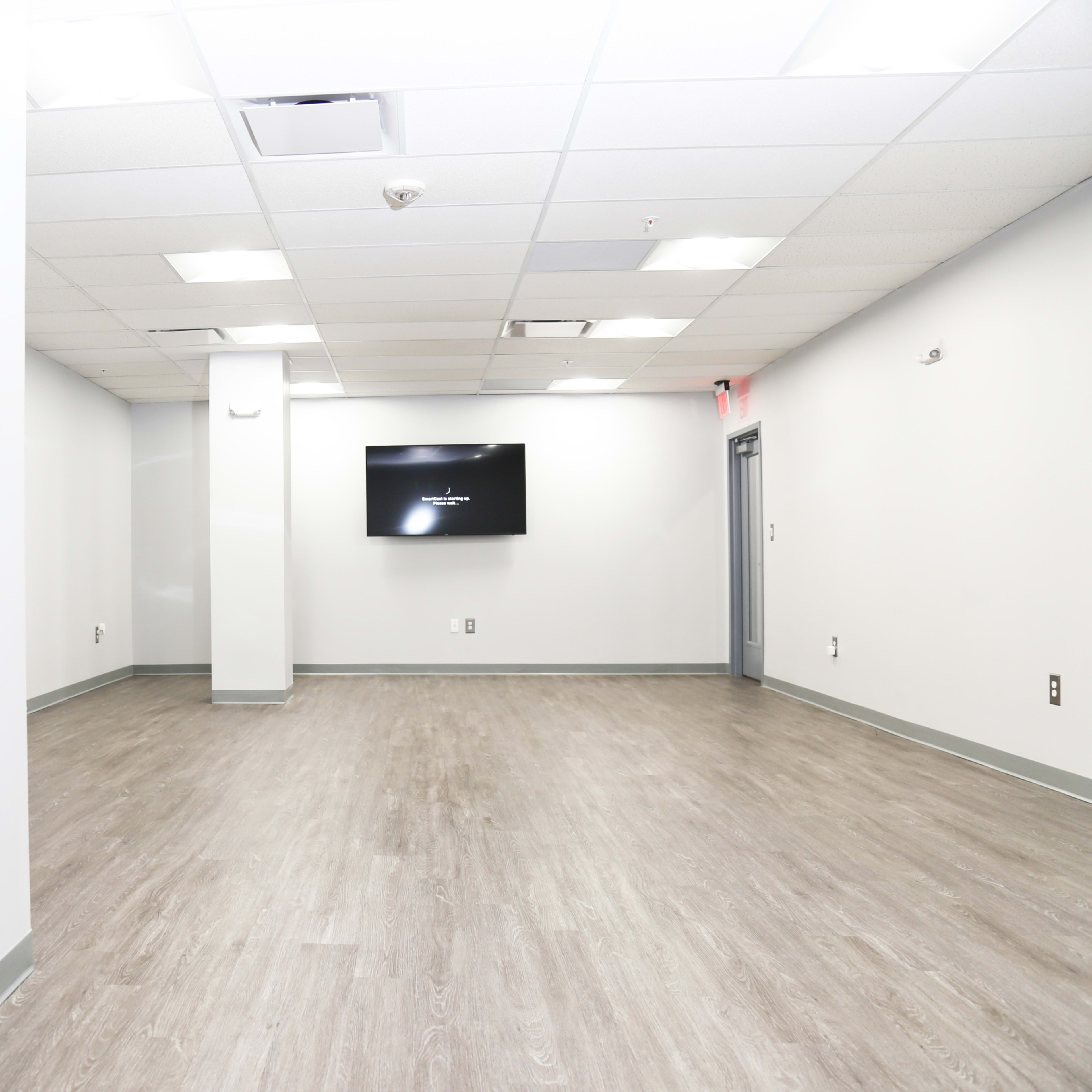 A spacious, empty office interior with light gray walls and a wooden floor, featuring a wall-mounted television.