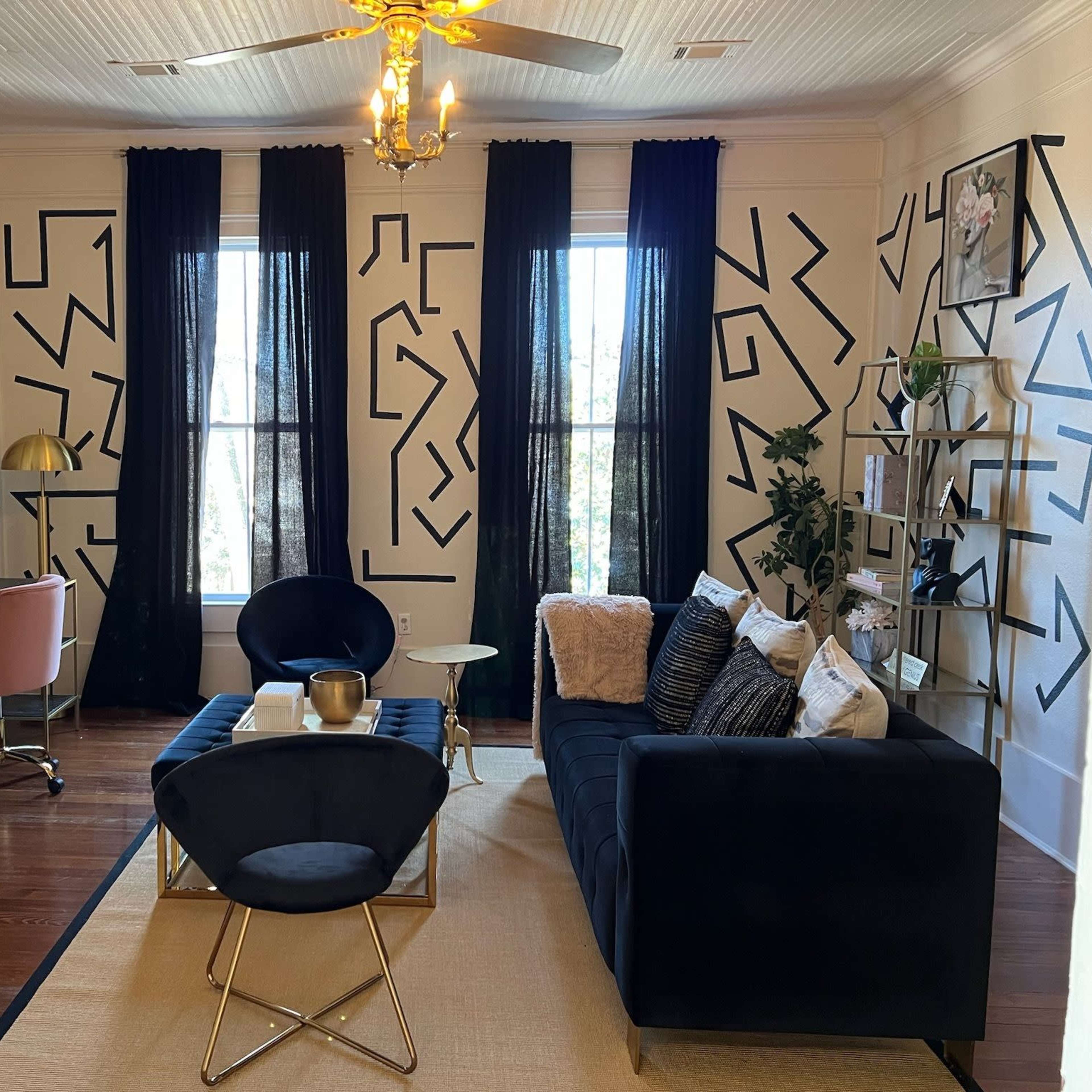 The living room features a dark blue sofa, a glass coffee table, and a desk with a pink chair, all set against walls adorned with black geometric patterns and large windows draped in dark curtains.