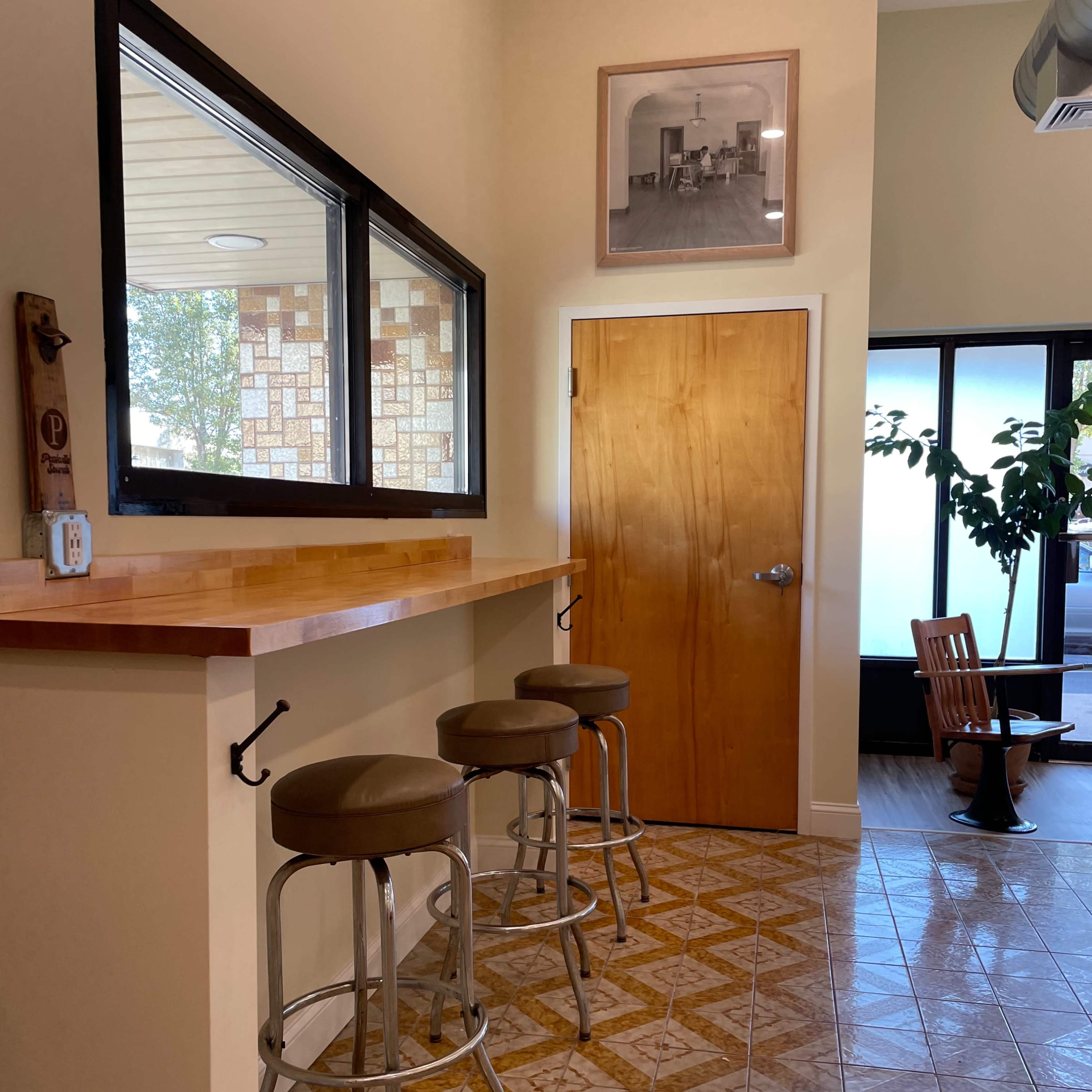 A bar area with three stools, a wooden countertop, and a door leading to another room, all set against a tiled floor and a framed photo on the wall.
