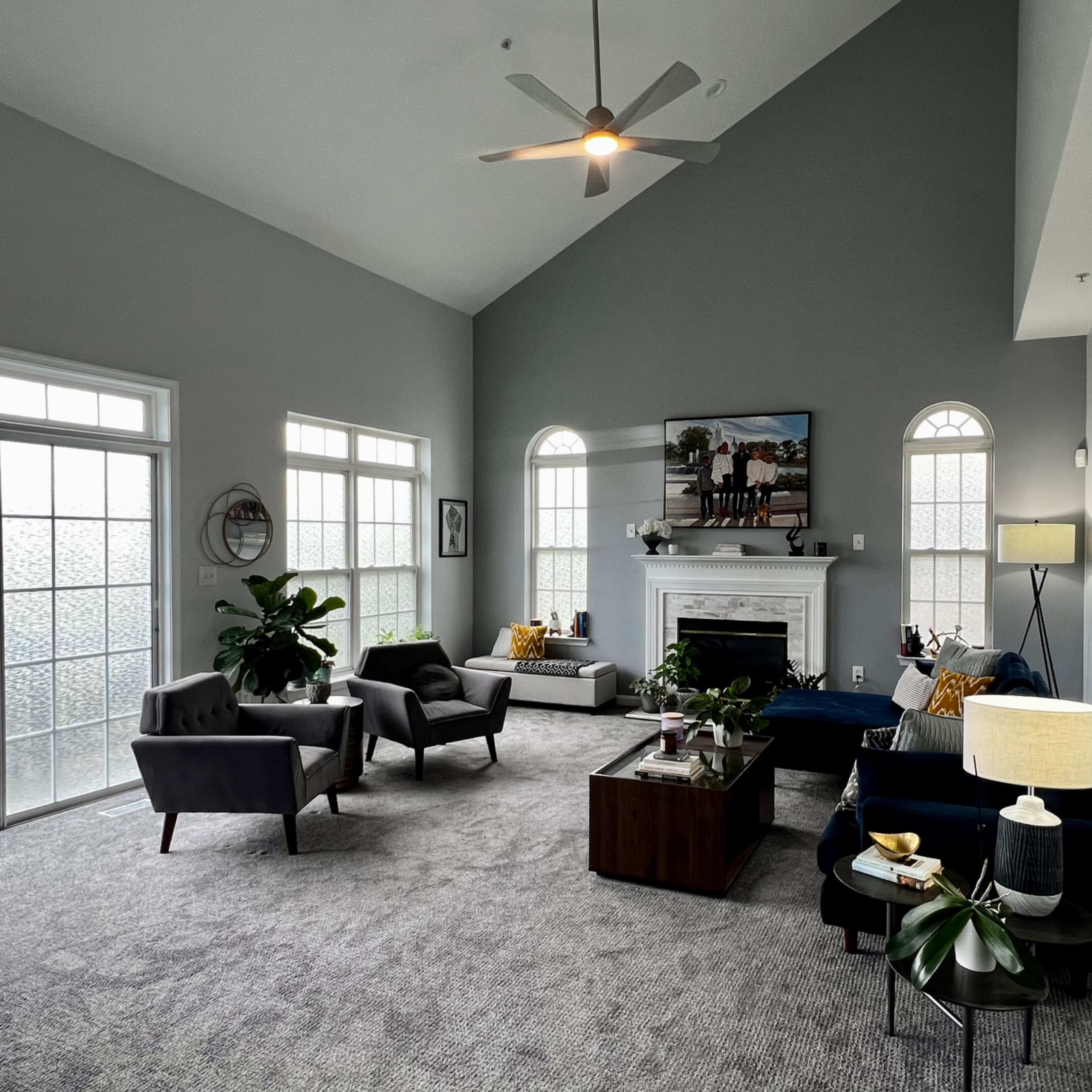 A spacious living room featuring a high ceiling, a fireplace, large windows, and modern furniture arranged around a coffee table.